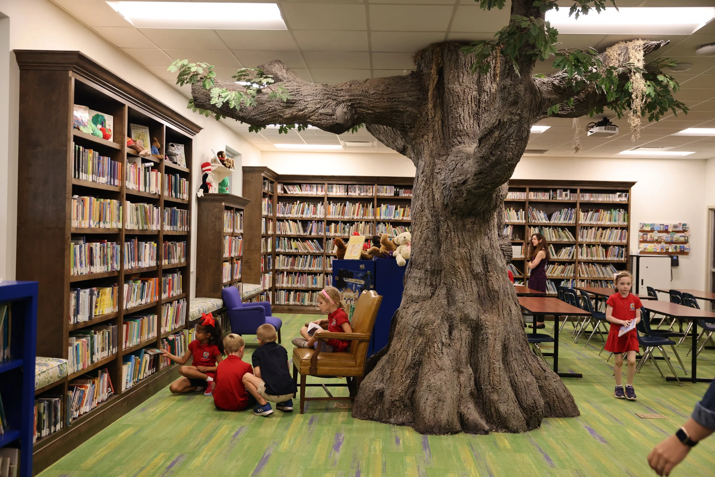 A large indoor space with a giant fake tree in the center, surrounded by children reading and sitting on chairs and there are bookshelves filled with books in the background.