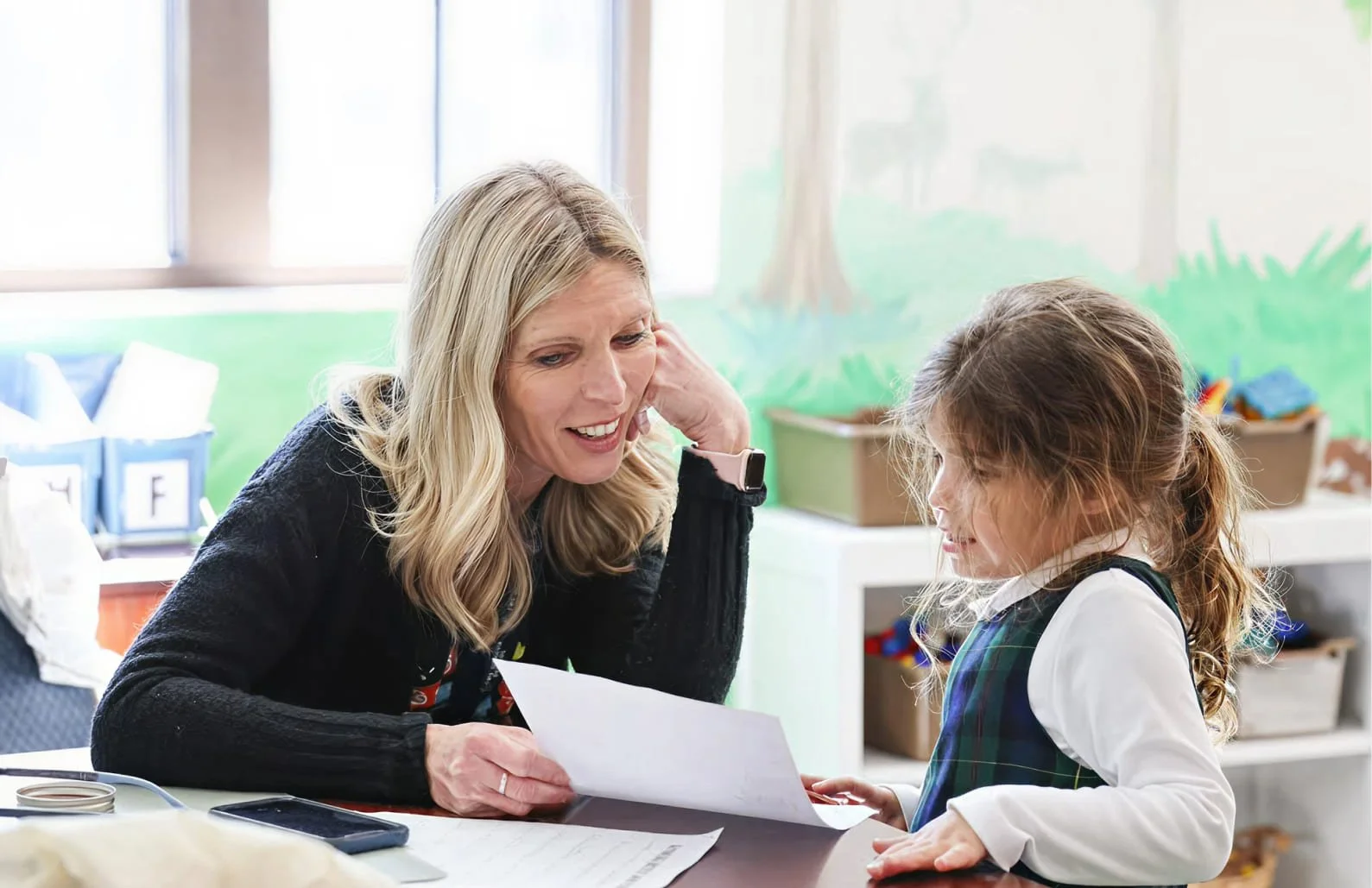 A teacher and a young girl sitting at a table, looking at a piece of paper together in a classroom setting with educational decor in the background.