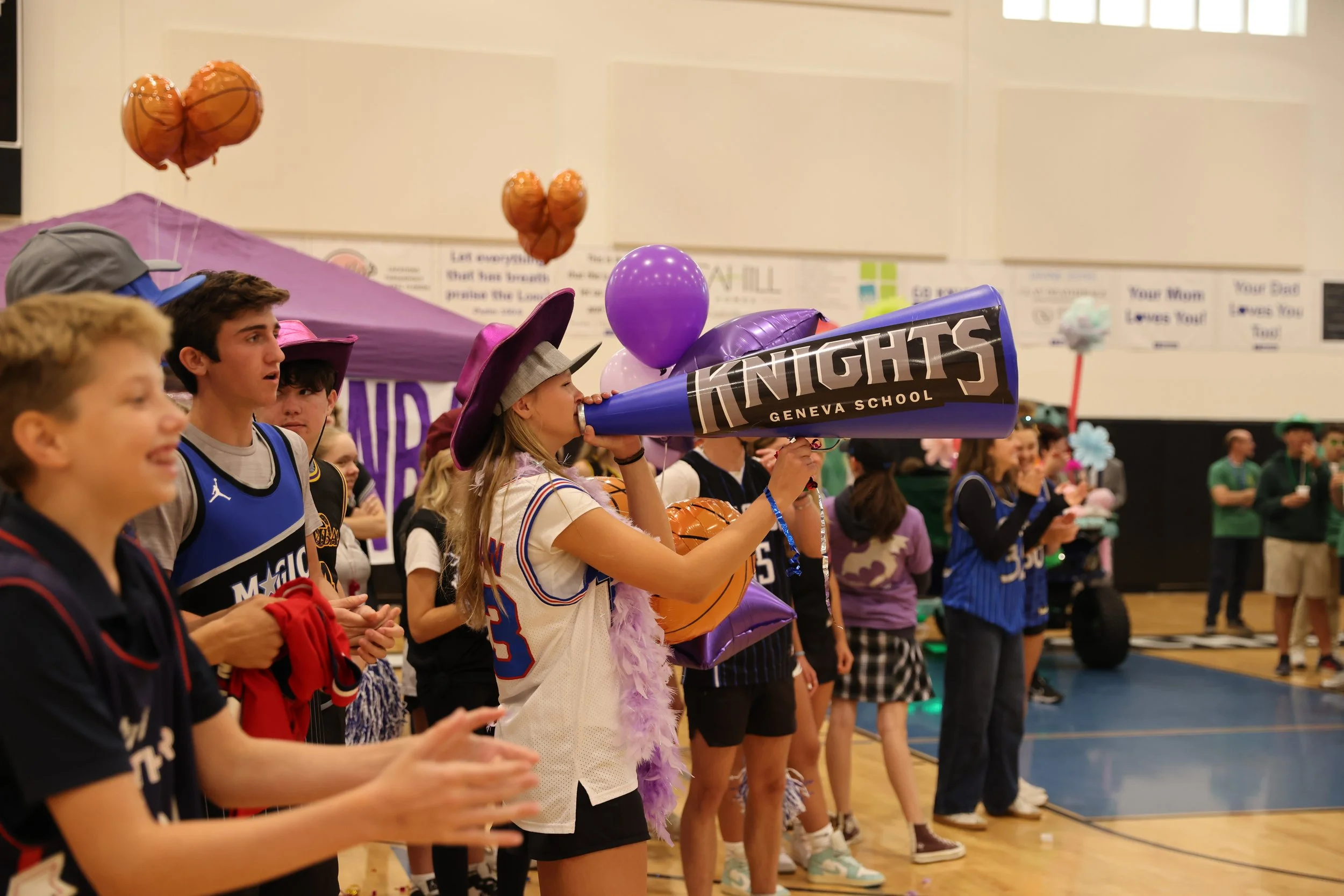 Students at a school event, some dressed in sports jerseys and others in costumes, lining up in a gymnasium, with balloons and banners in the background.