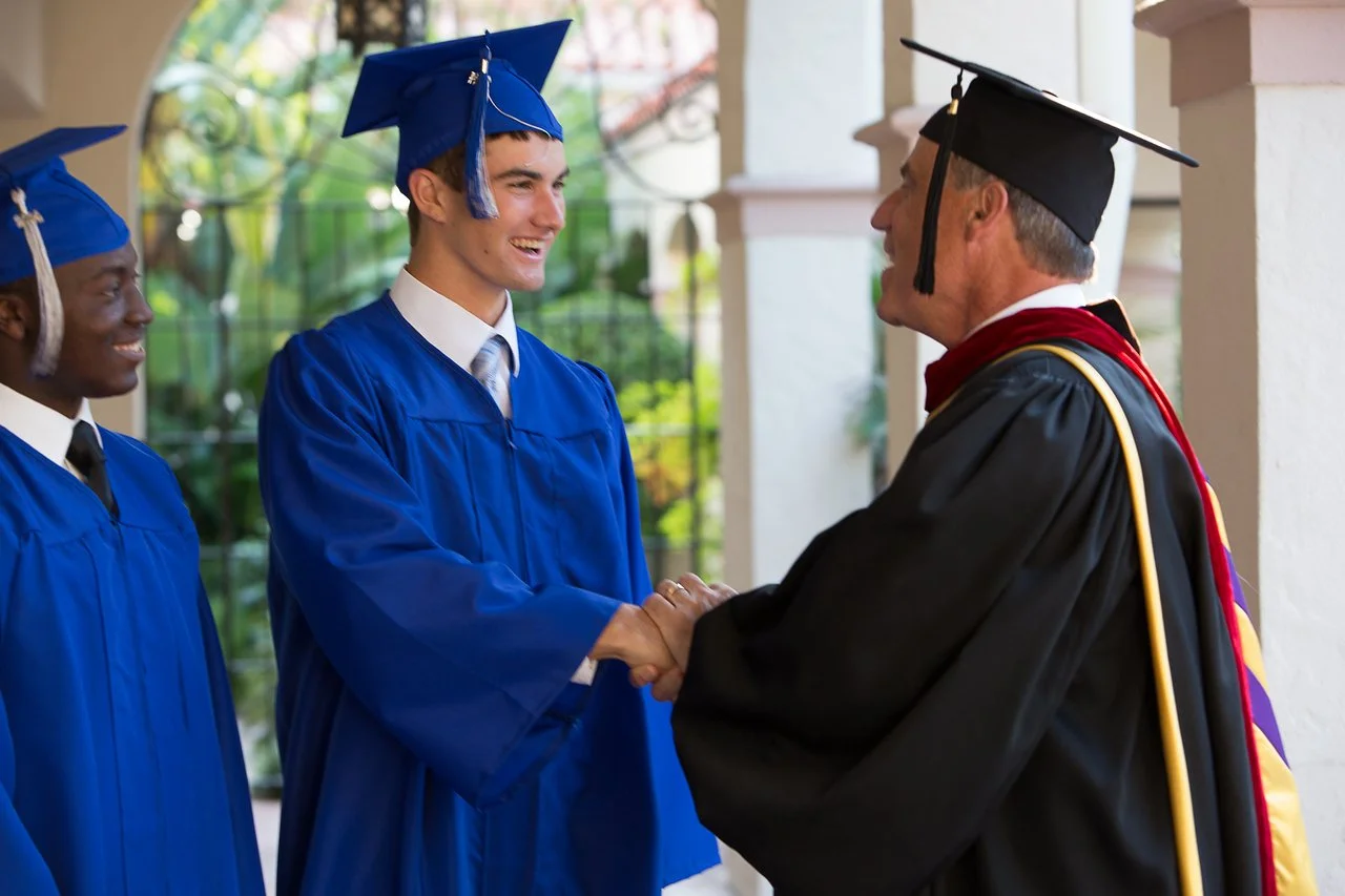 Graduate in blue cap and gown shaking hands with faculty member in black academic regalia, other graduates looking on.