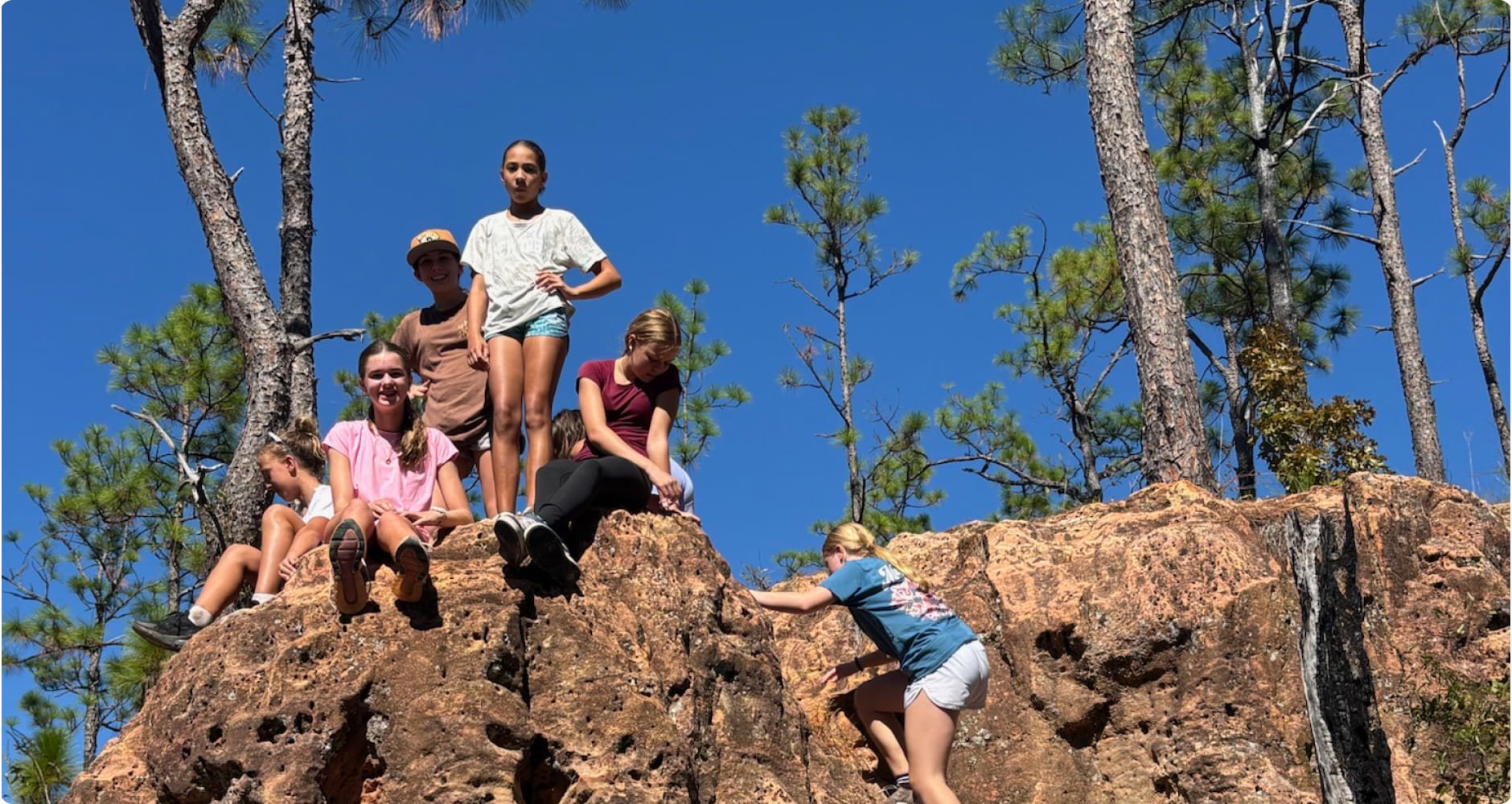 Group of children playing and climbing on rocky terrain in a wooded outdoor area under a clear blue sky.