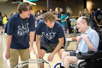 Two young men, wearing matching navy blue T-shirts, are talking to an older man in a wheelchair. The setting appears to be a busy hospital or community event.