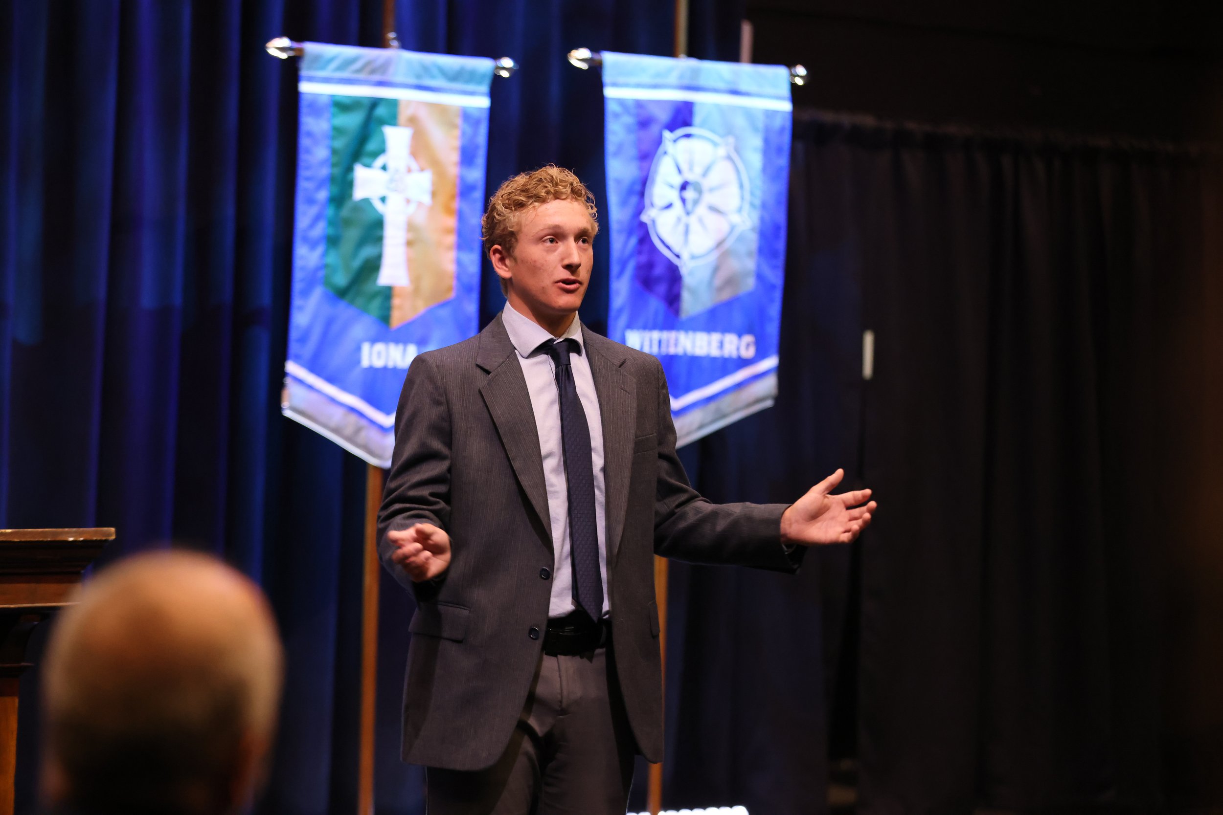 A young man in a gray suit and tie speaking or giving a presentation on stage, with banners featuring crosses and symbols in the background.
