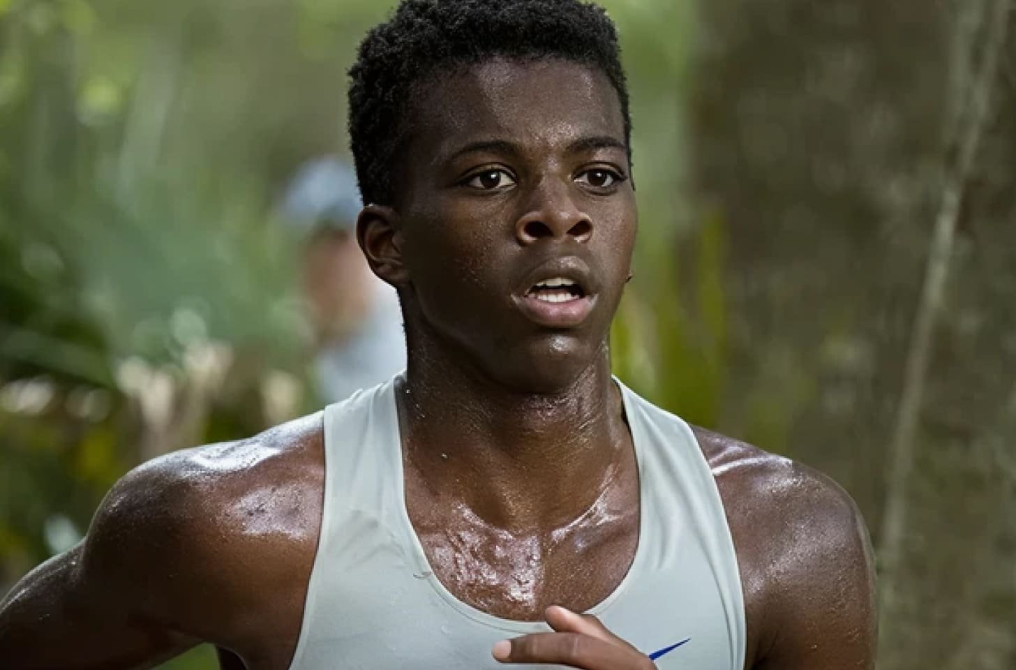 A young male runner with dark skin and short curly hair on a trail in a forest, sweating and wearing a sleeveless athletic shirt.