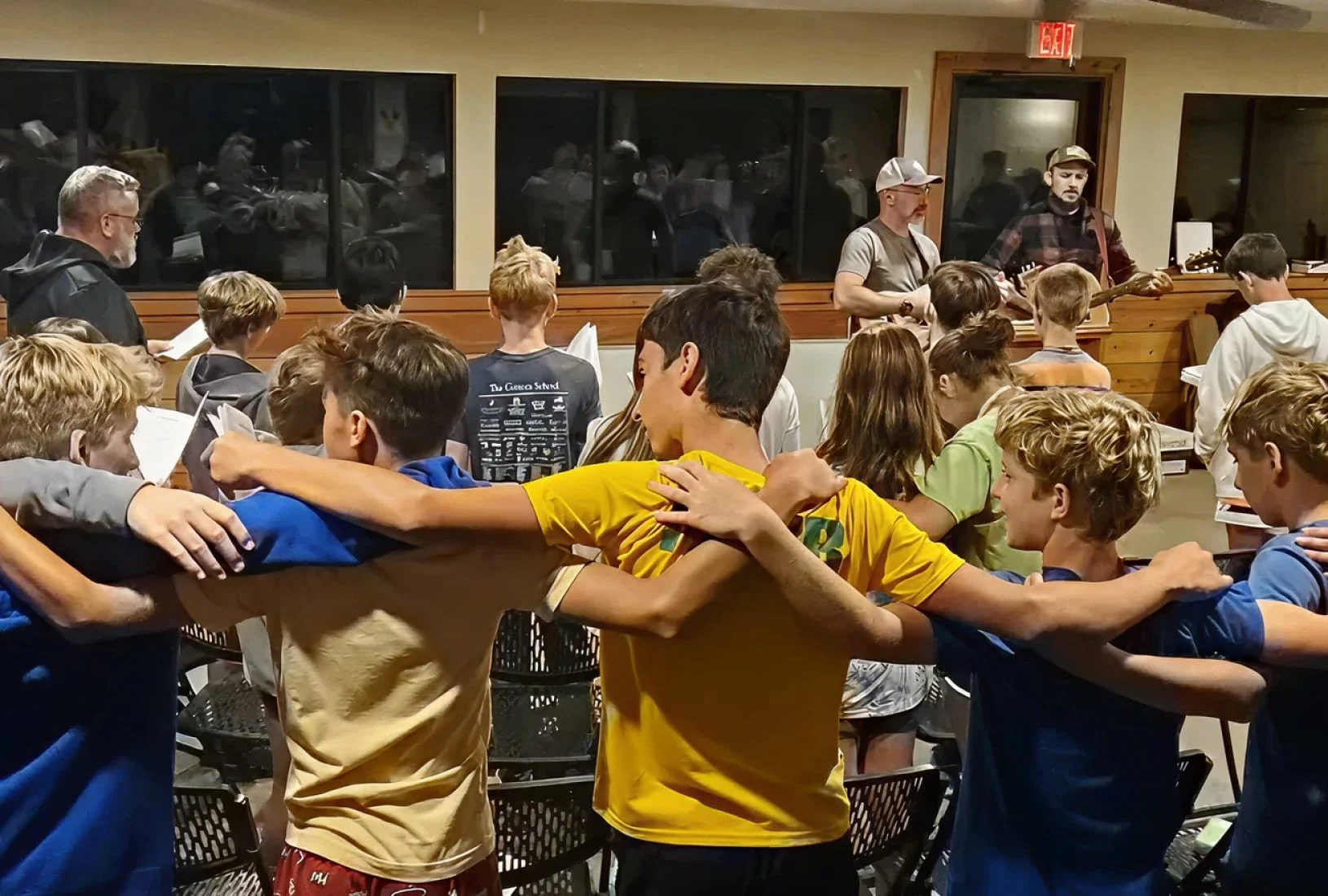 A group of students in a circle with arms around each other, participating in a musical performance or practice, with two musicians playing guitars at the front in a room with large windows reflecting reflections of people.