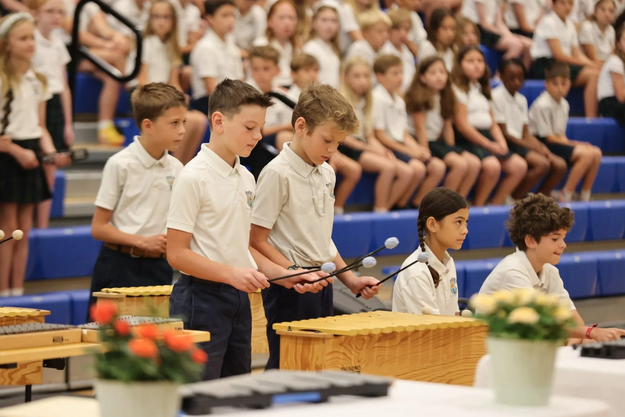Children wearing white uniforms playing xylophones at a school event, with other students seated behind them.