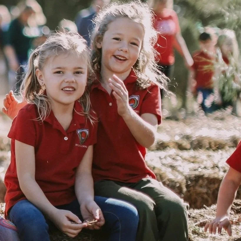 Two young girls in red Geneva school uniforms sitting outdoors, smiling at the camera, with other children in similar outfits in the background.