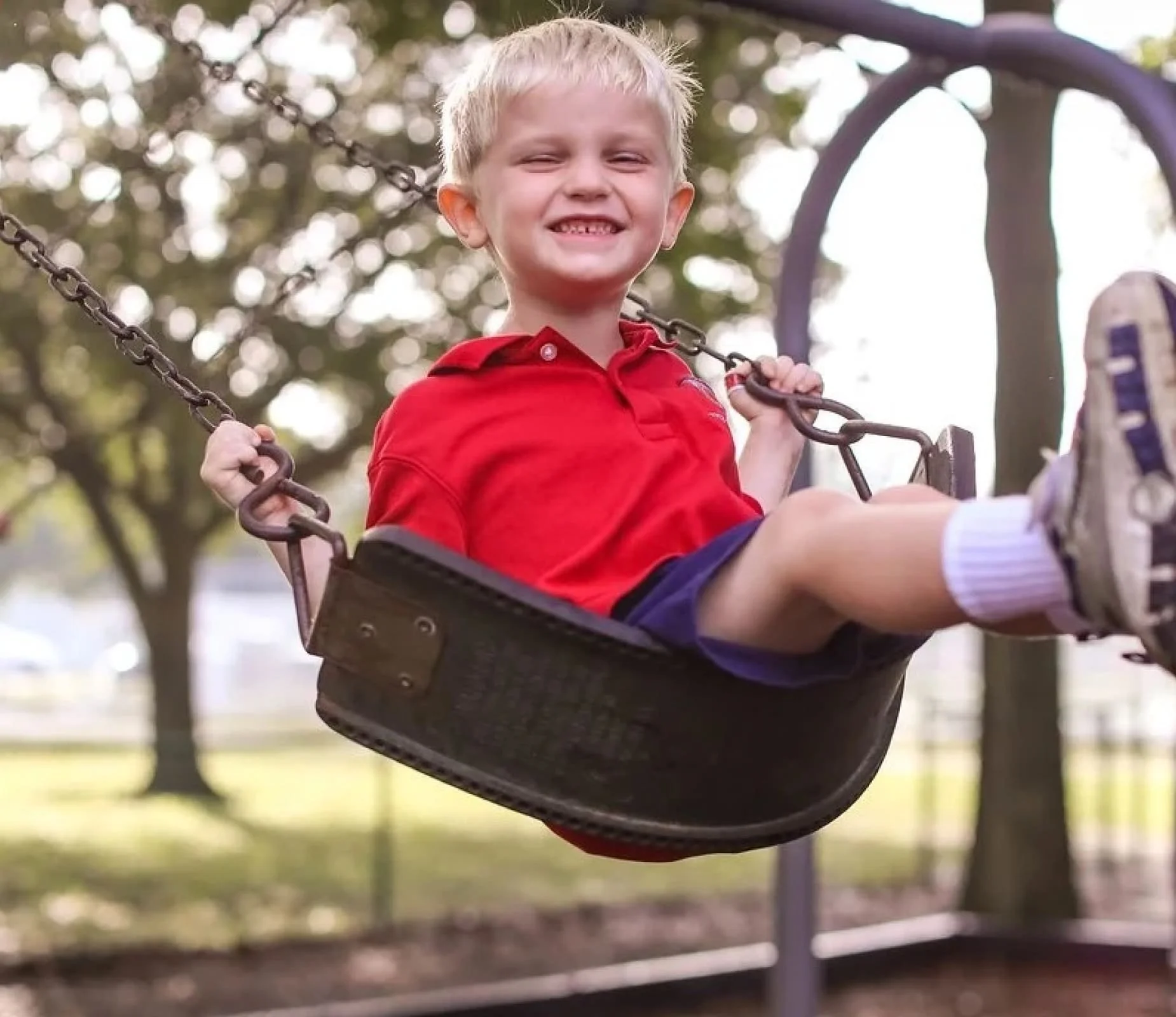 A young boy smiling as he sits on a playground swing, holding onto the chains, with trees and a park in the background.