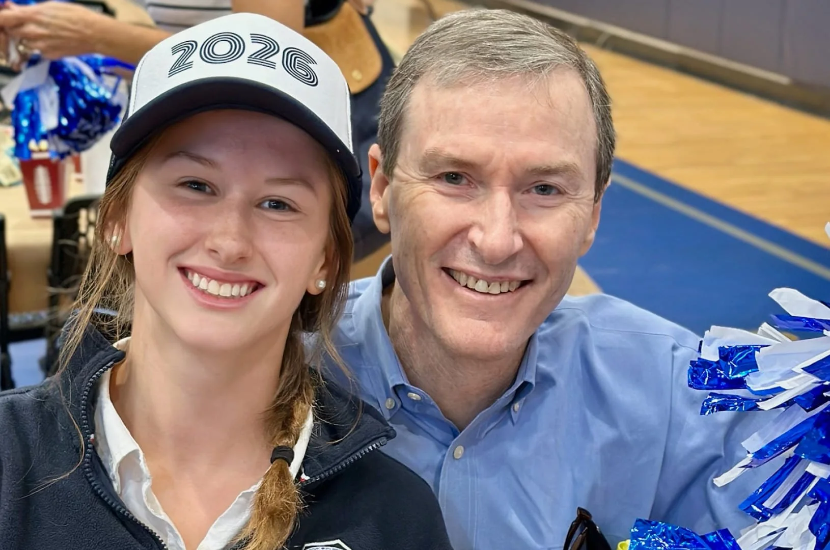 Photo of a girl wearing a white college cap with '2026' on it, and a man, both smiling for the camera indoors at a celebration or event.