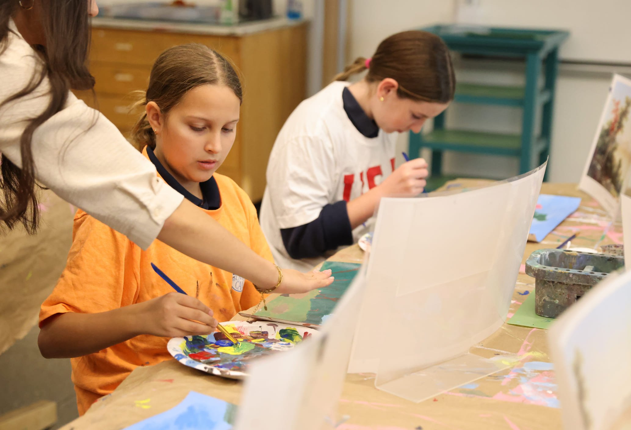 Two young girls and an woman painting at a table in an indoor art classroom, with art supplies visible in the background.
