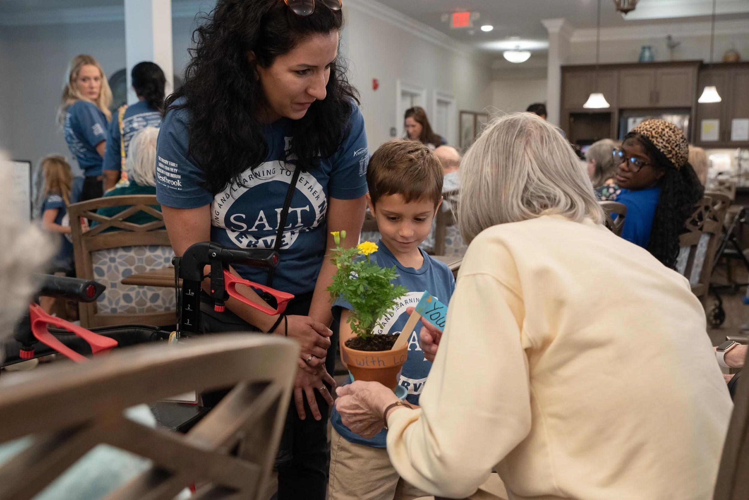 A woman and a young boy planting a small yellow flower together at an indoor event, with other people in the background.