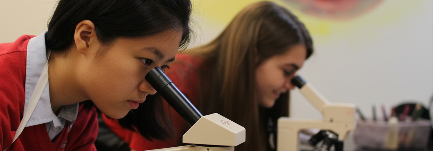 Two young women looking into microscopes in a classroom or laboratory setting.