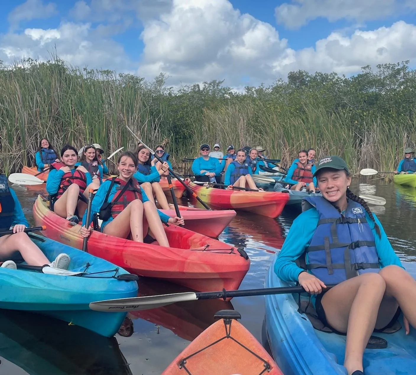A group of young people kayaking on a calm waterway surrounded by tall grasses and trees, with some people smiling and holding paddles, under a partly cloudy sky.