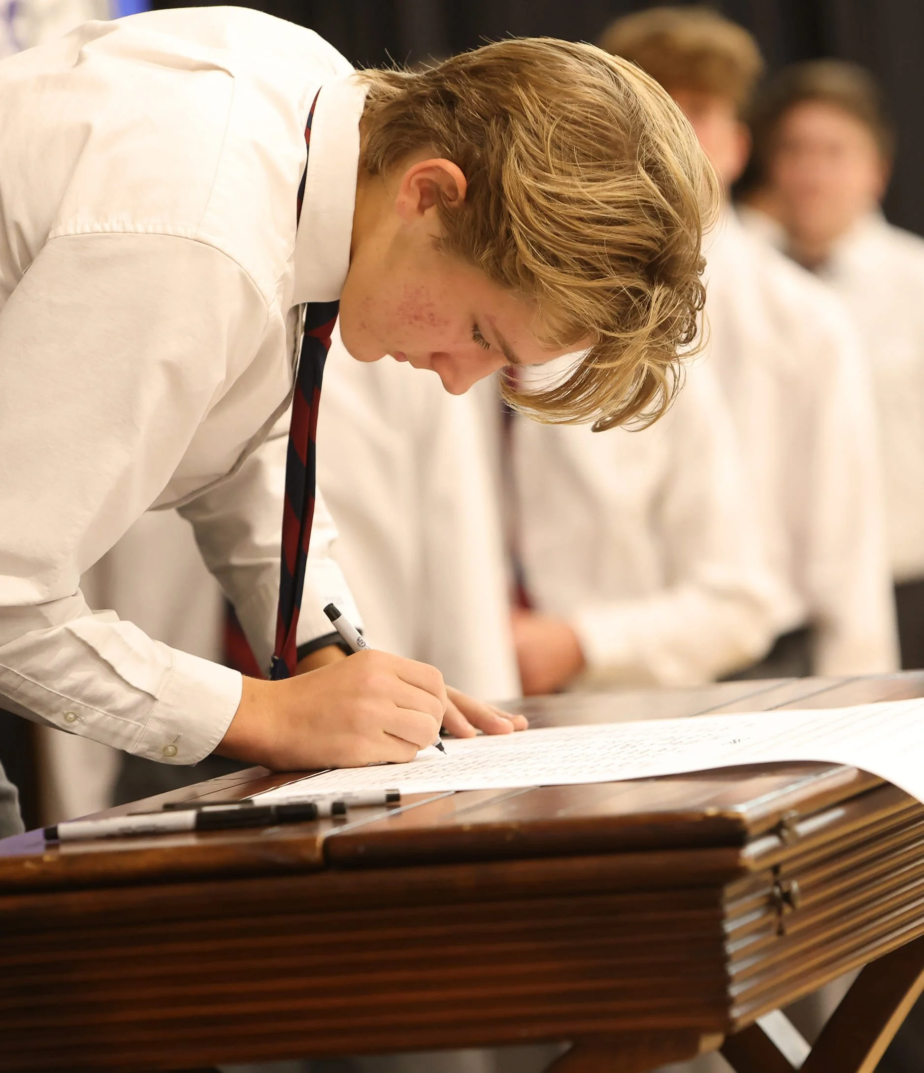 A student with wavy blond hair and wearing a white shirt signs a document on a wooden table, with other people standing behind him.