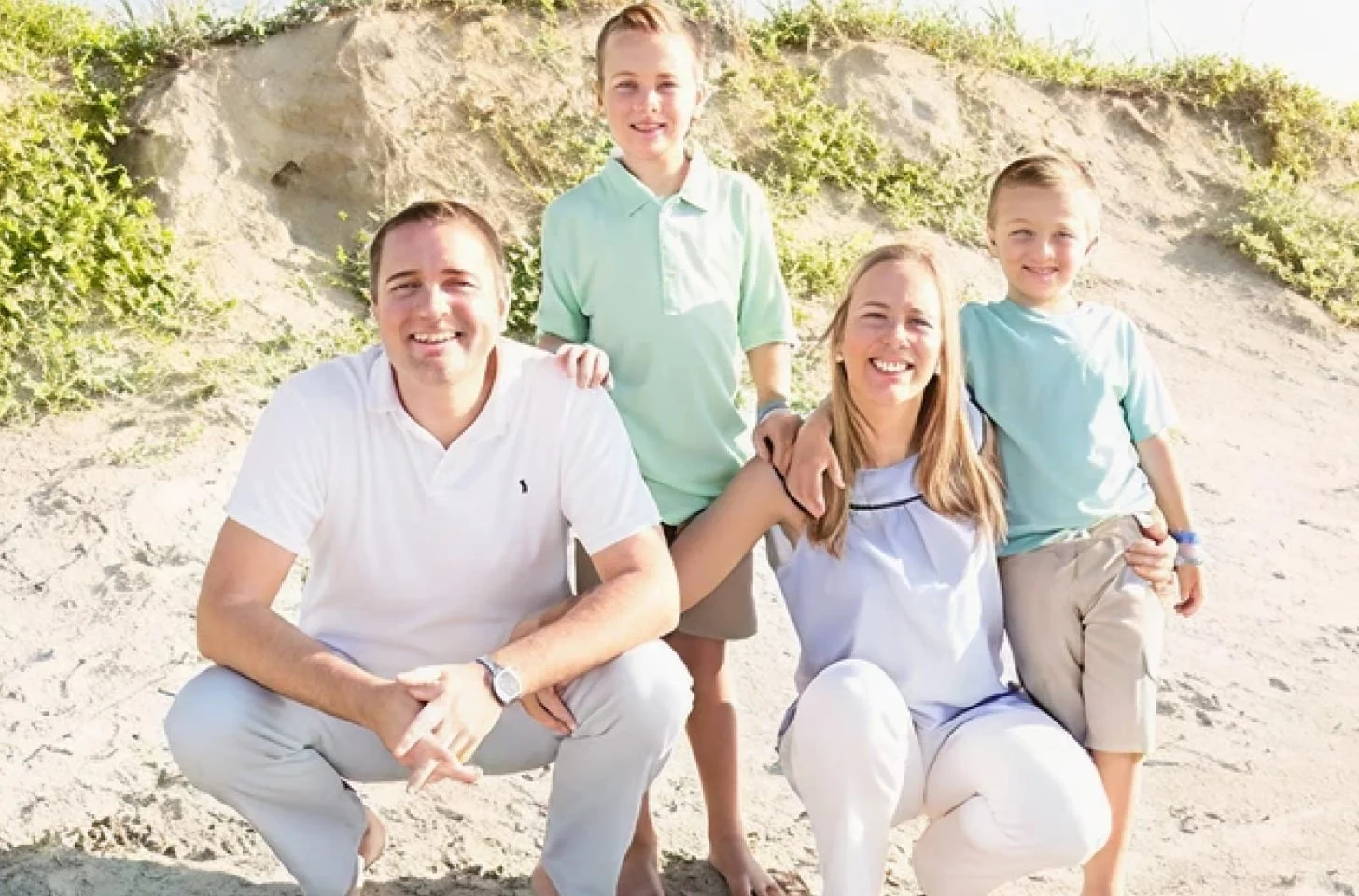 A family of five posing on a sandy beach with green bushes and rocky formations in the background.