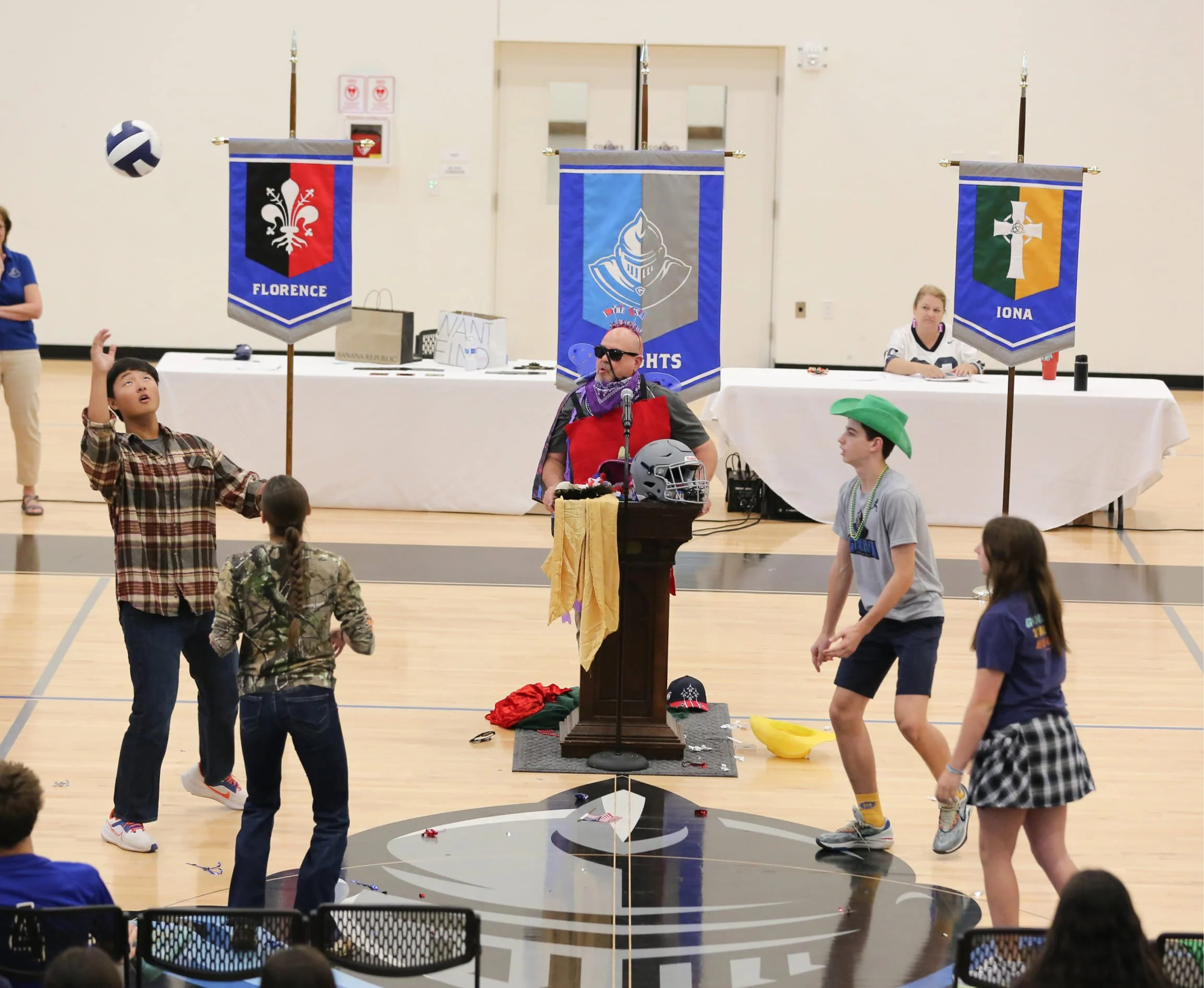 Children and adults in a gymnasium participating in a game or activity around a wooden table with banners hanging above. One banner reads 'FLORENCE,' another depicts a knight's helmet with the words 'THE KNIGHTS,' and the third banner says 'IONA' with a cross and shield. The scene appears lively with children reaching towards a volleyball, and there is a magician or entertainer with sunglasses and a purple bandana leading the activity.