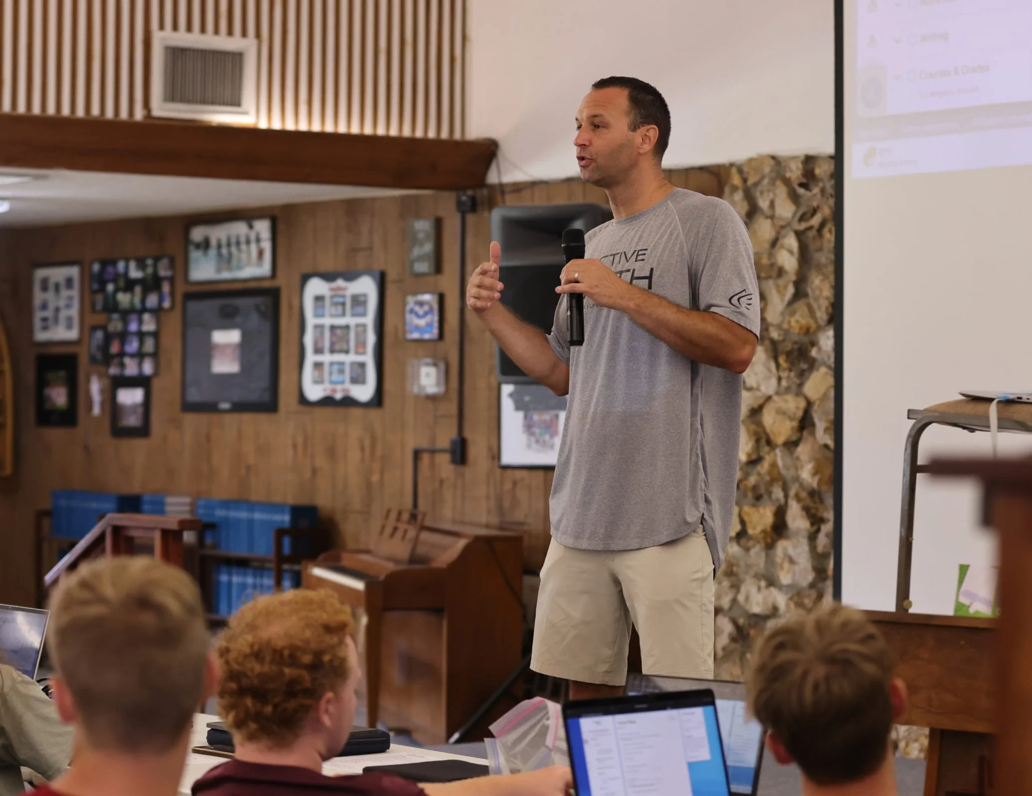 A teacher giving a presentation in a room with wood-paneled walls and a stone fireplace, speaking into a microphone while an audience of children or teenagers listens, some working on laptops.