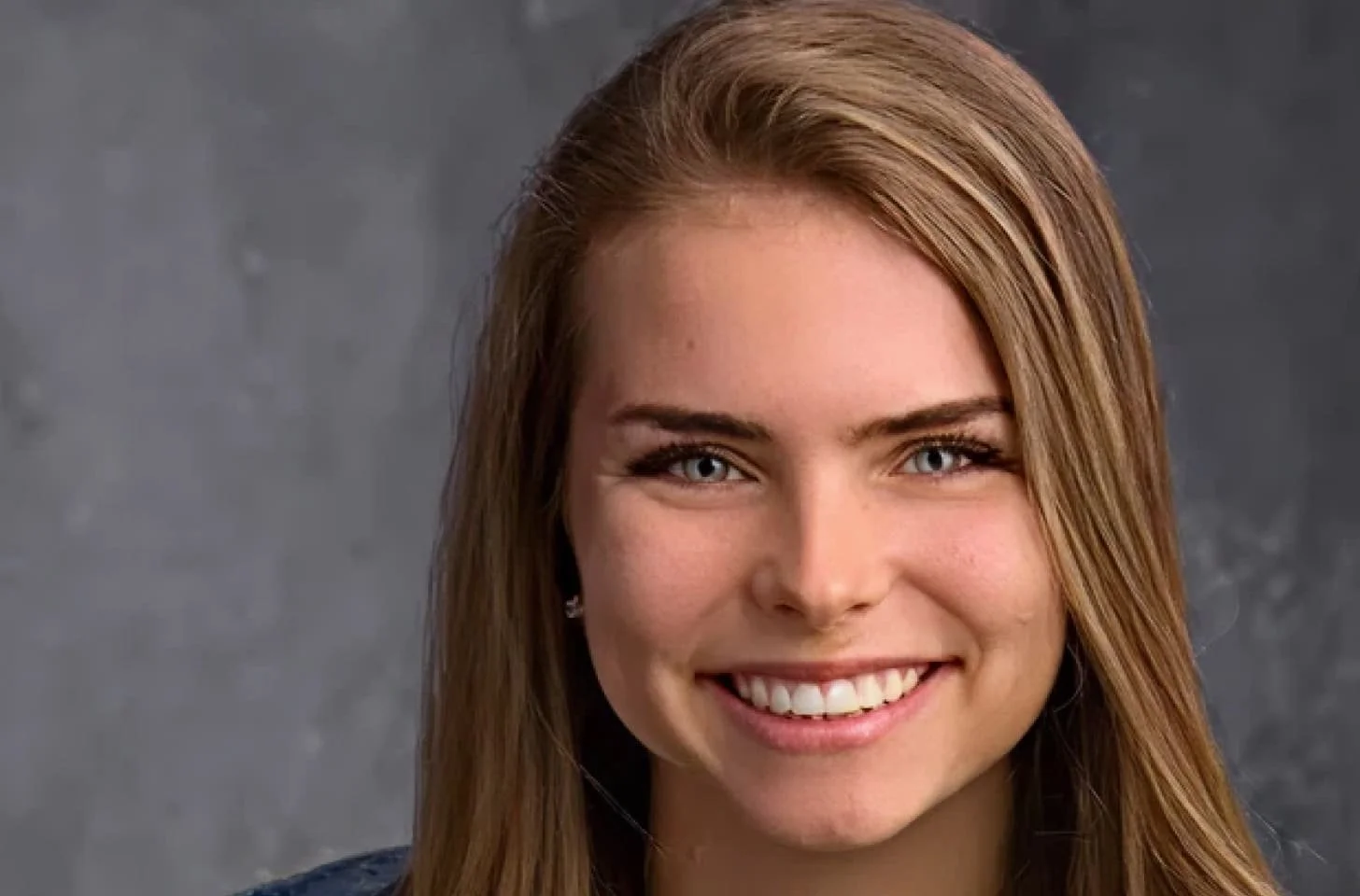 Close-up portrait of a young woman with long light brown hair, blue eyes, and a bright smile, wearing small earrings, against a gray background.