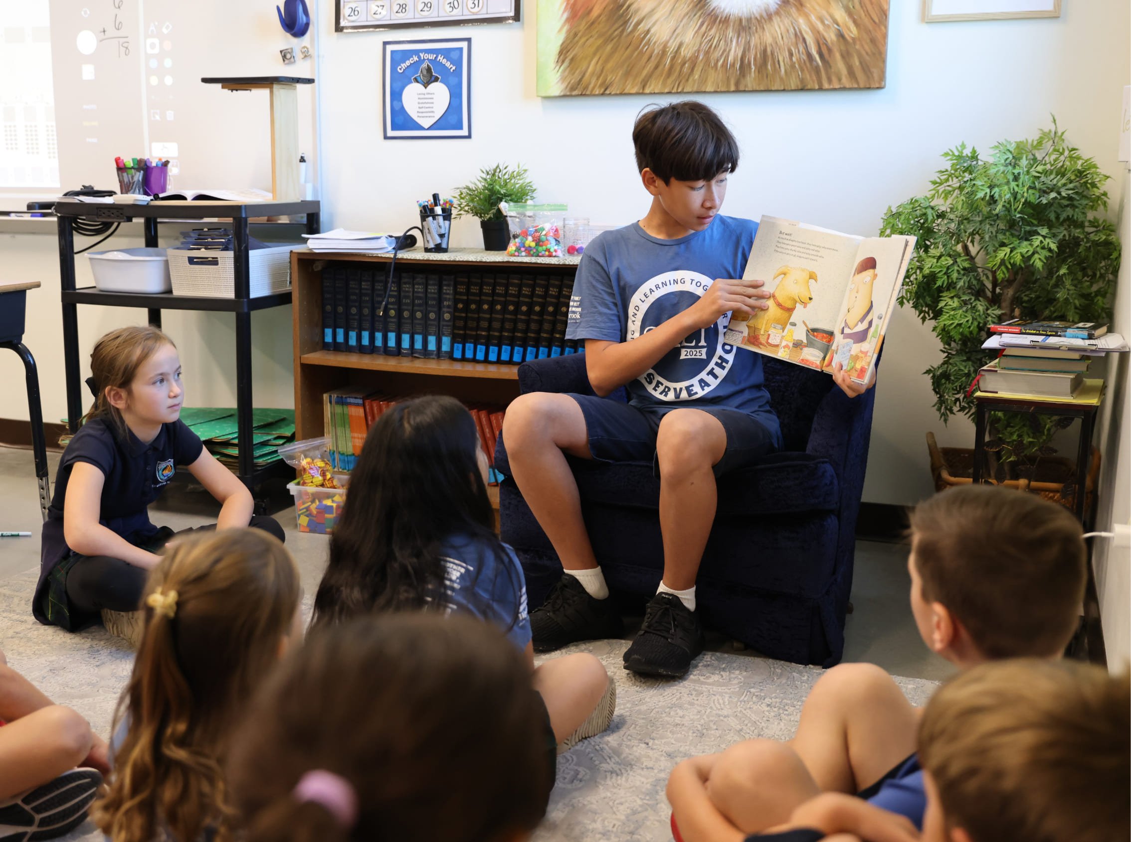 A group of children sitting on the floor listening to a boy reading a picture book in a classroom.