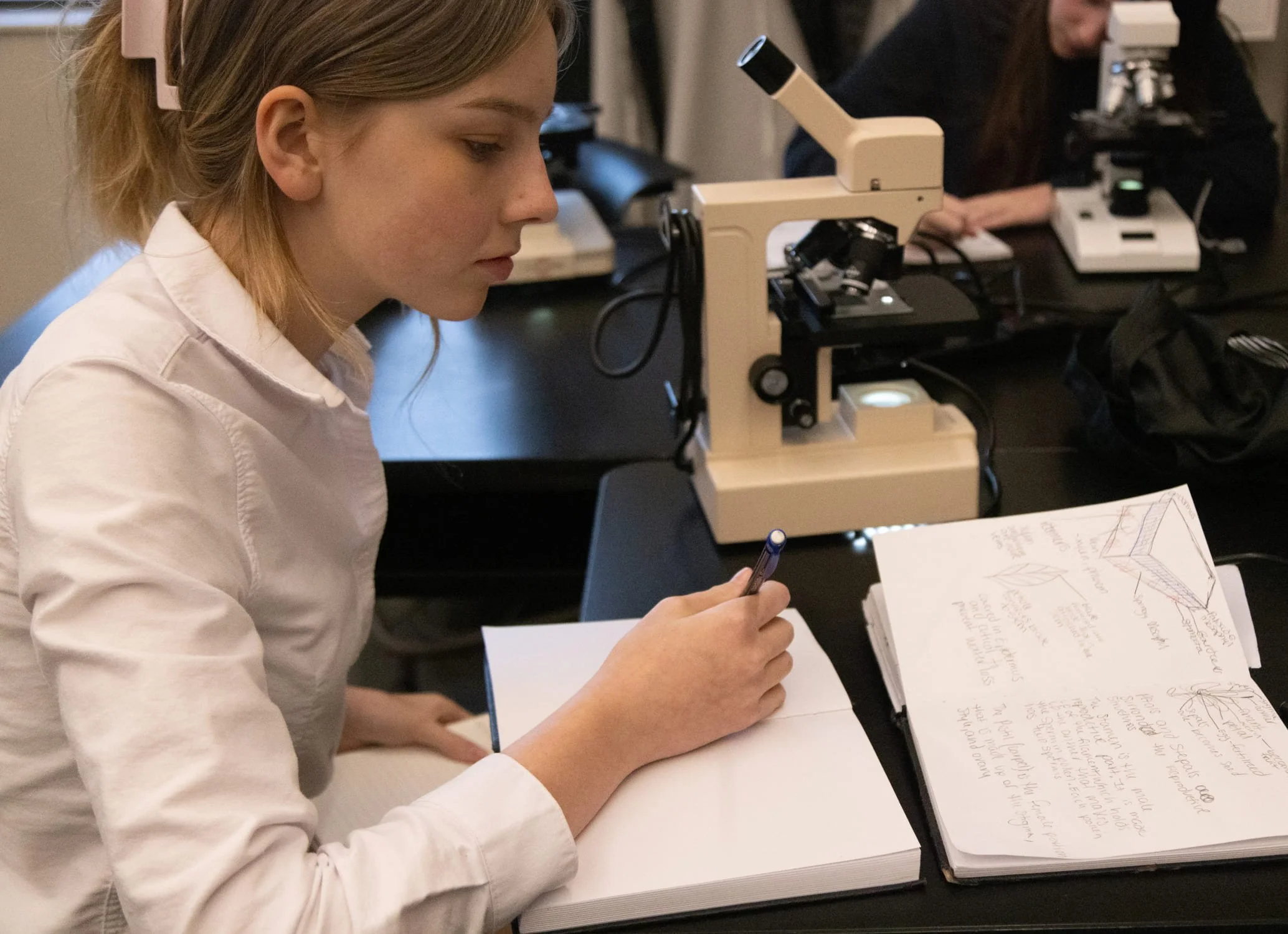 A student with long hair tied back and wearing a white shirt is sitting at a desk, looking through a notebook filled with handwritten notes.