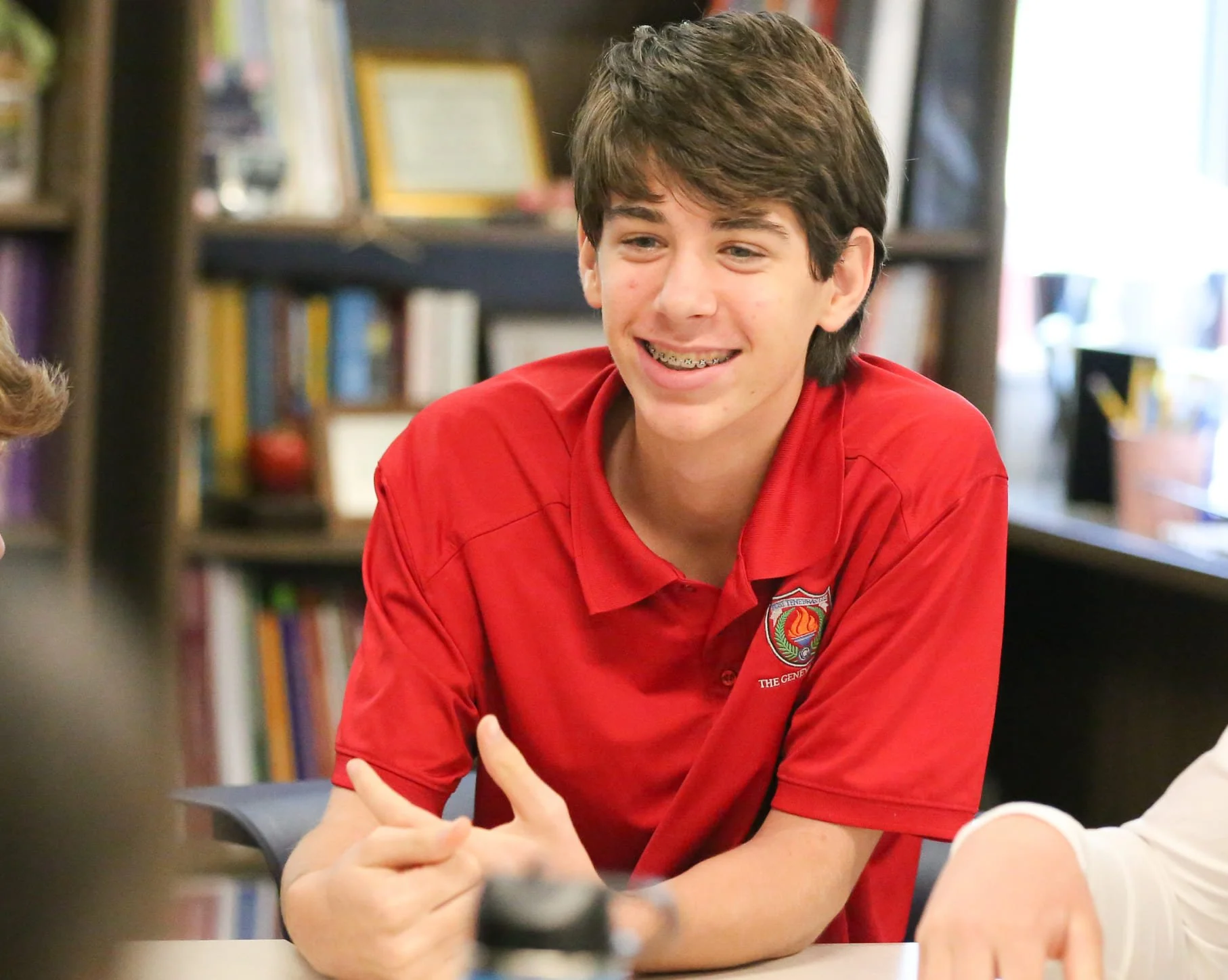 A teenage boy with brown hair wearing a red collared shirt with a Geneva logo, smiling and talking with someone at a table in a room with bookshelves and framed pictures in the background.