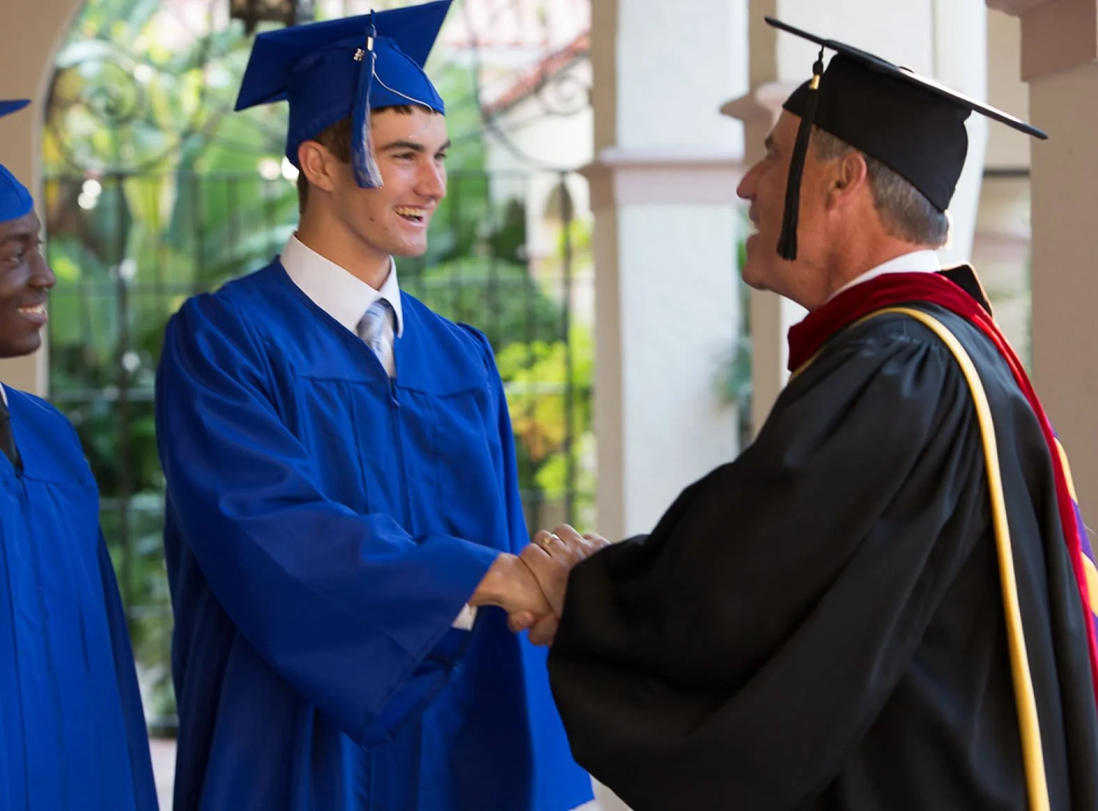 A young man in a blue graduation gown and cap shaking hands with a man in academic regalia during a graduation ceremony.