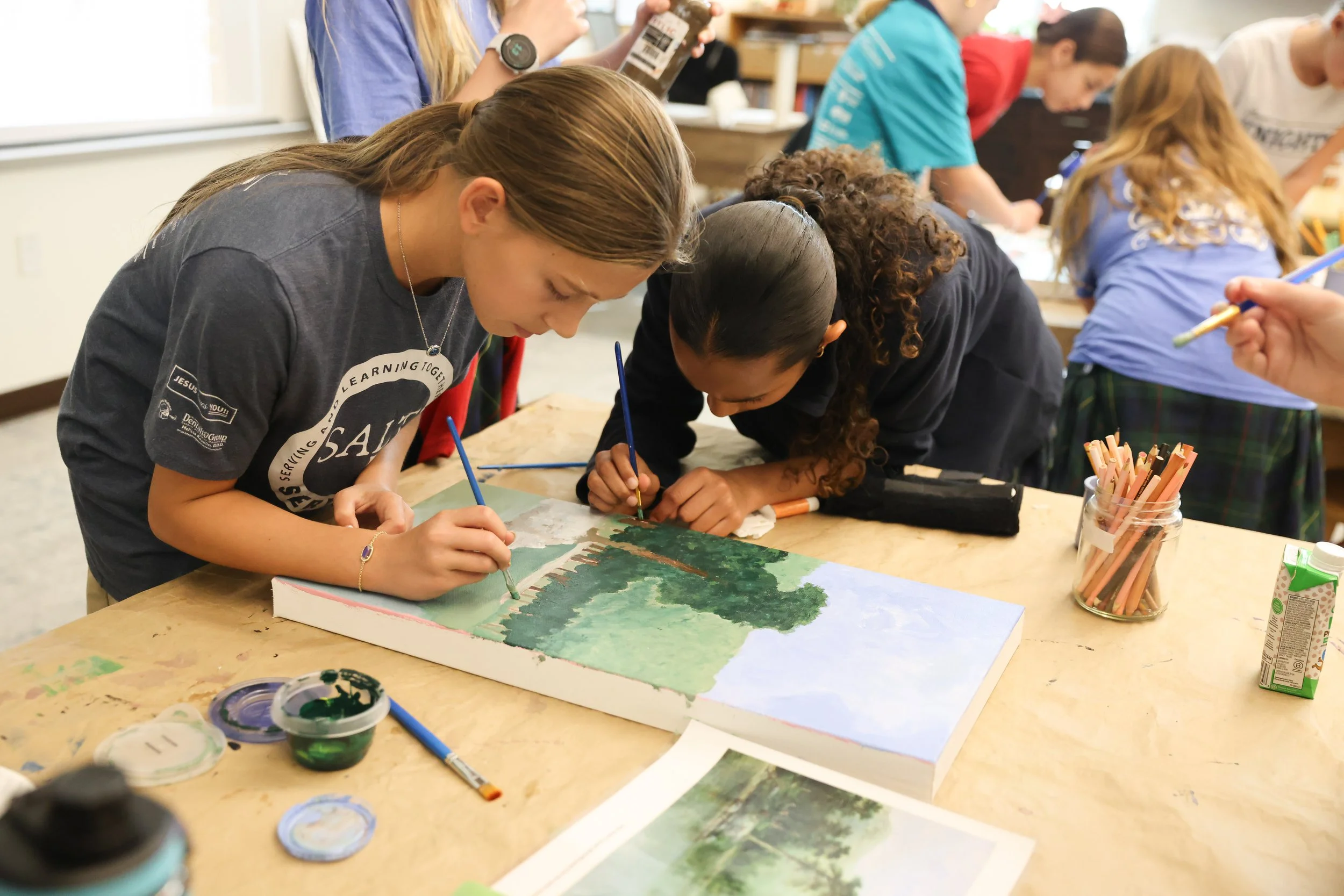 Students in a classroom are working on a painting project at a table. They are focused on their artwork, with some painting trees and nature scenes. Art supplies like brushes, paint, and colored pencils are on the table.