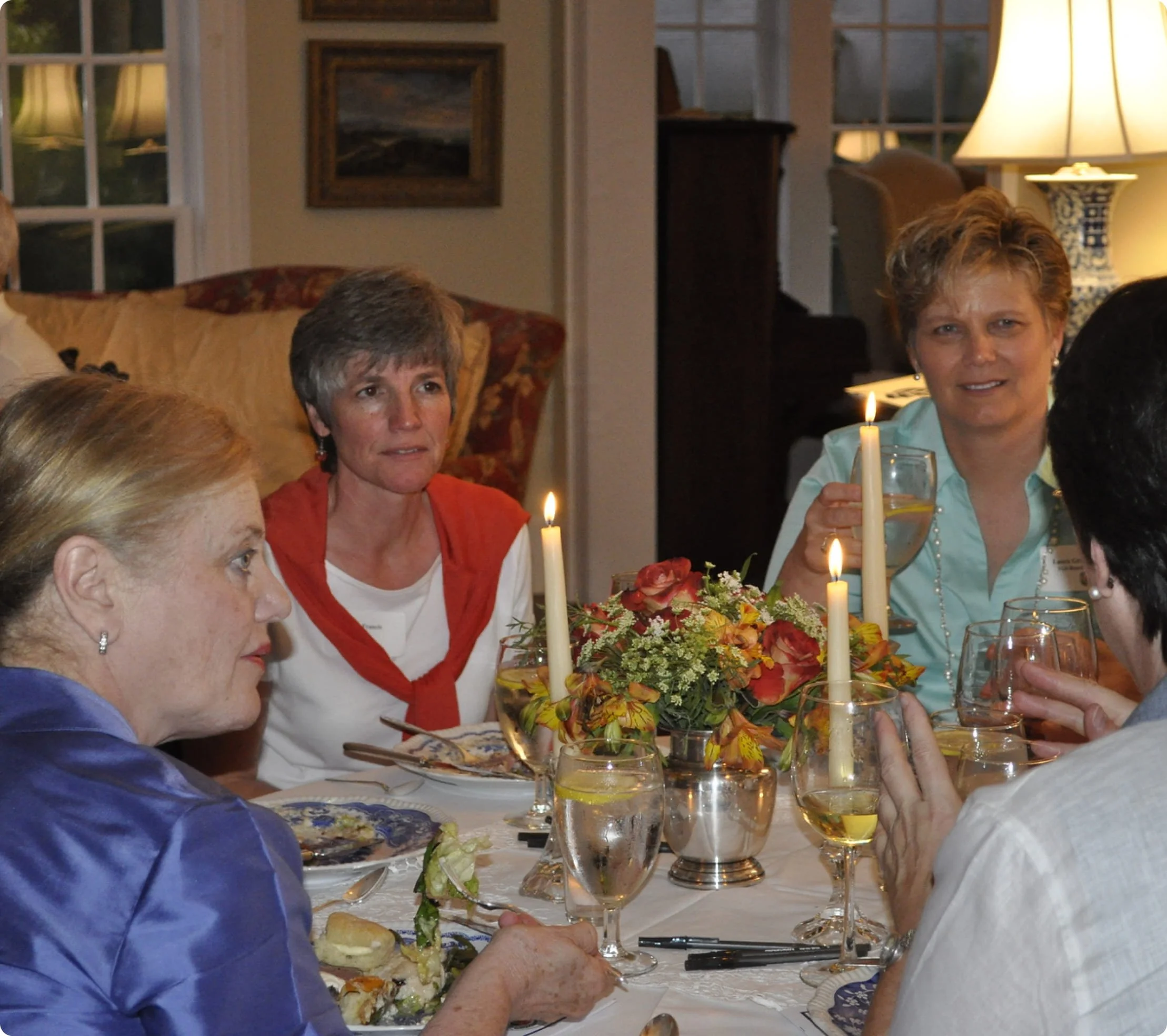 People dining at a formal dinner table decorated with candles and floral arrangements in a cozy, well-lit room.