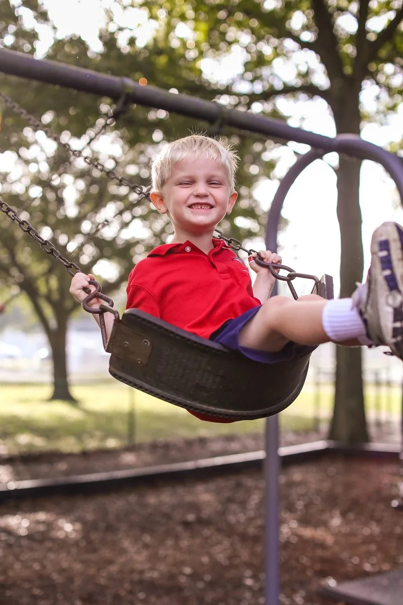Young boy in a red shirt smiling on a swing at a park with trees in the background.