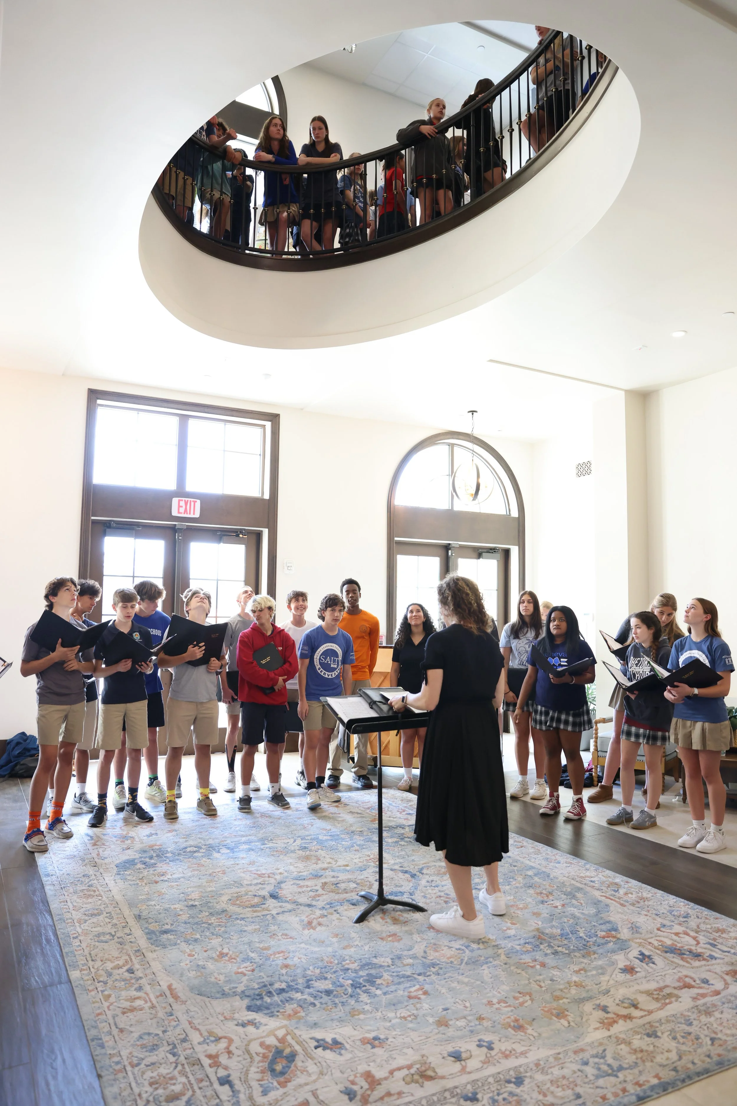 A choir of young students singing, directed by a female choir conductor, inside a well-lit room with large windows and a decorative area rug.