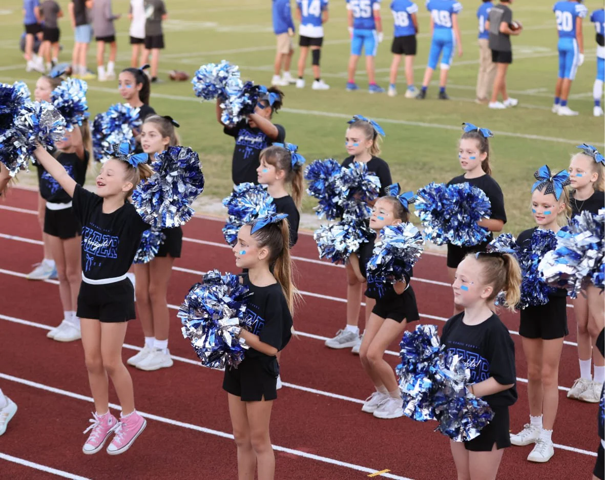 A group of young cheerleaders in black shirts and shorts, with blue bows in their hair, holding blue and silver pom-poms, standing on a red running track during a sports event. In the background, football players in blue uniforms and spectators are on the field.