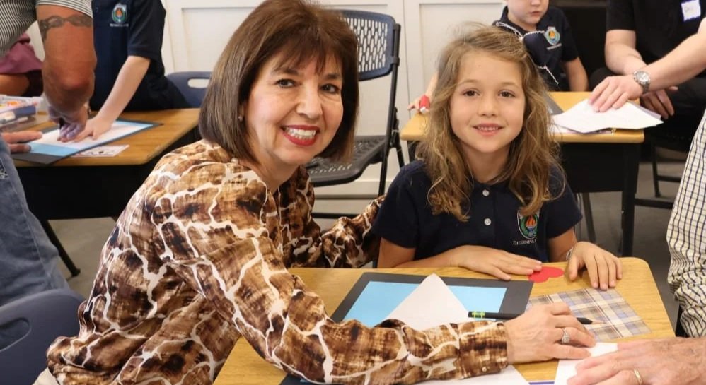 A teacher smiling and sitting at a table with a young girl in a classroom or meeting room.