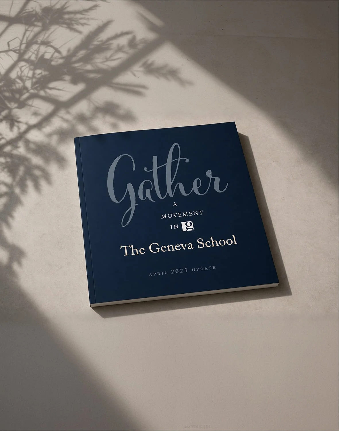 A blue book titled 'Gather: A Movement in The Geneva School, April 2023 Update' placed on a light-colored surface with shadows of a plant's leaves cast on the surface and partly on the book.