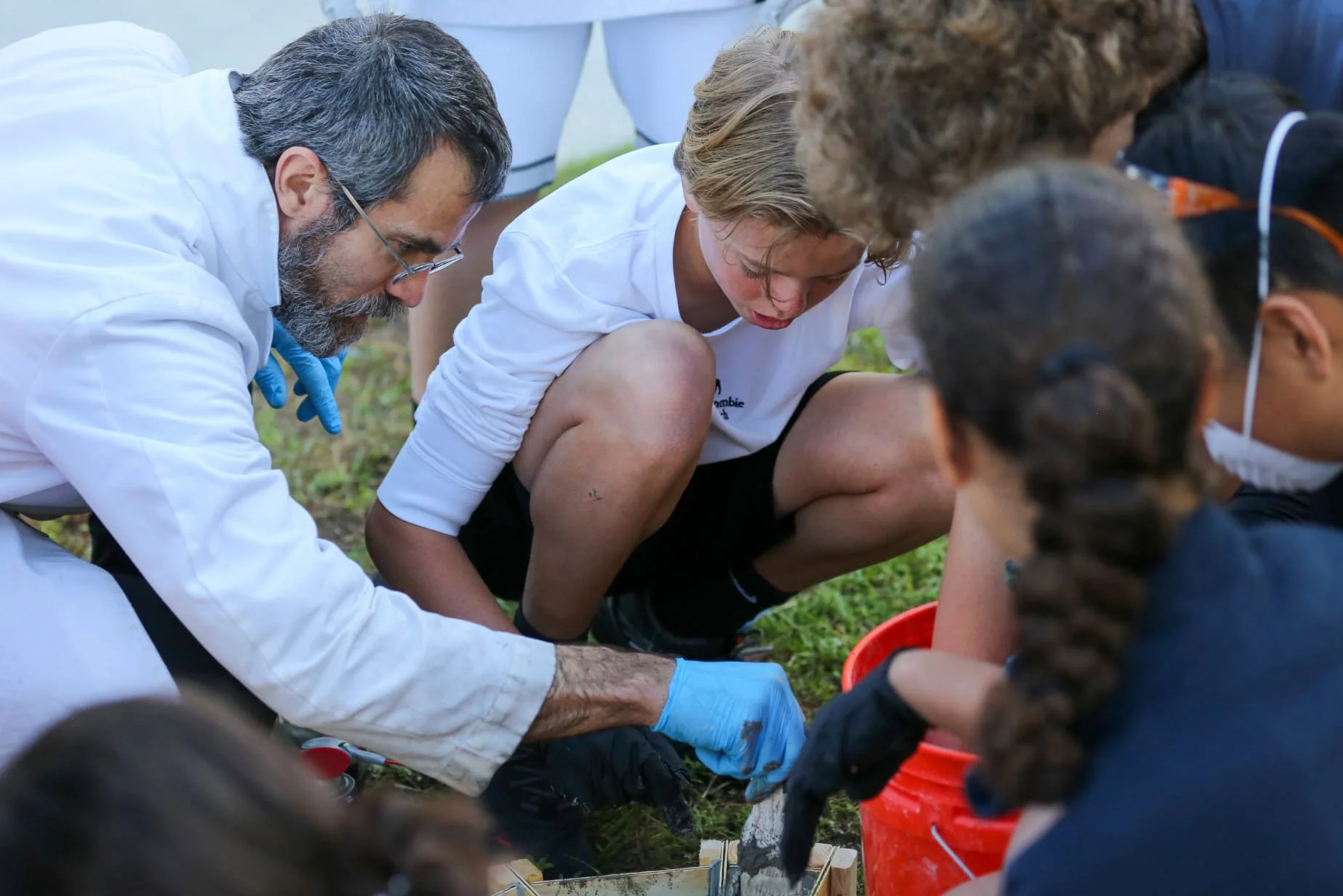 Group of children and an adult scientist working together outdoors on a project, with the children wearing face masks and the scientist wearing gloves.