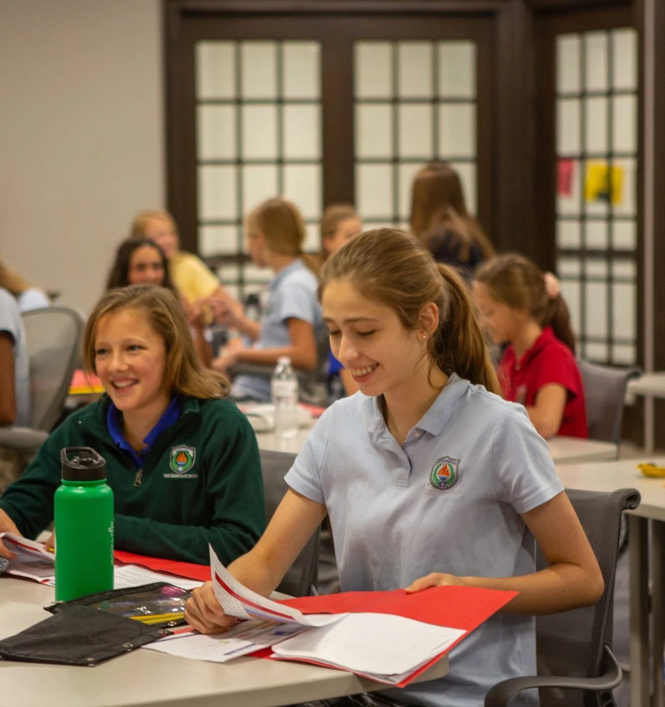 Young girls in school uniforms sitting at tables, smiling and interacting in a classroom or conference setting.