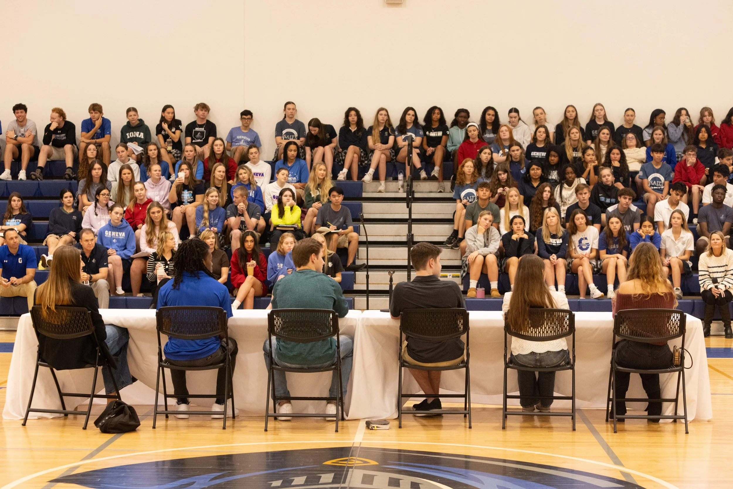 A large group of young people, mostly girls, sitting on bleachers in a gymnasium, facing forward and listening to a panel of officials seated at a long table covered with a white tablecloth in front of them.