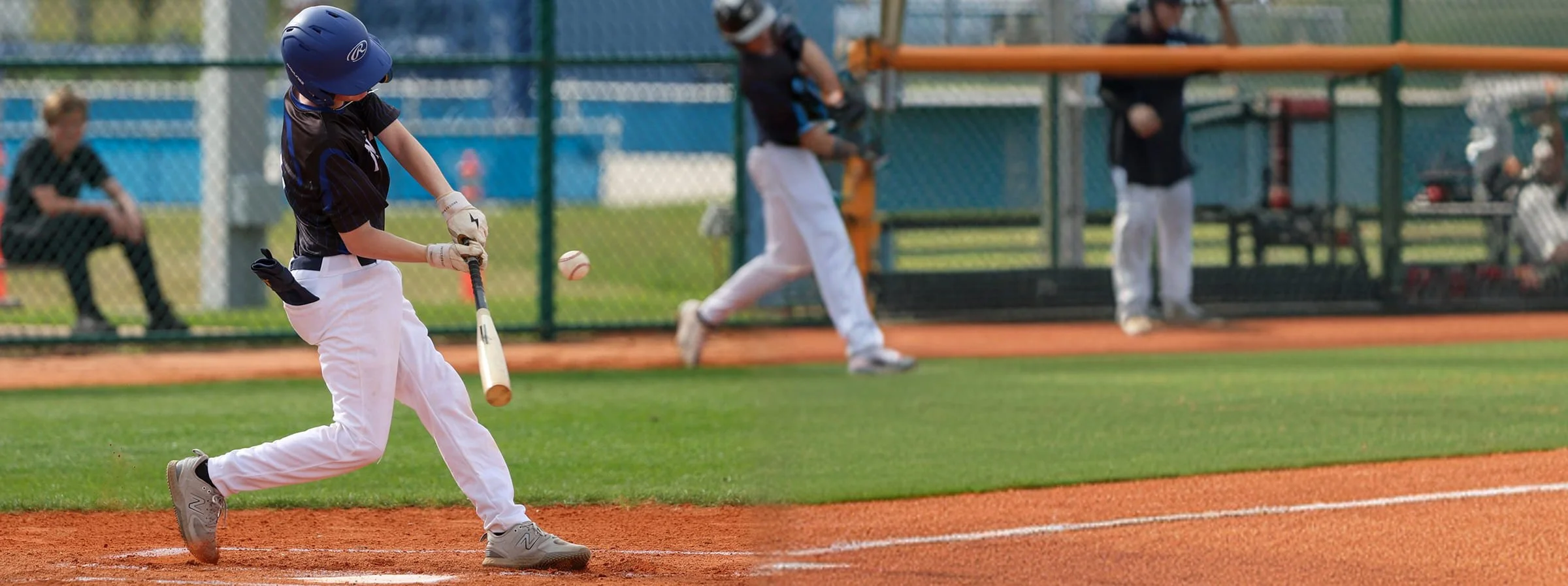 A young baseball player in a black shirt, white pants, and a blue helmet, is running towards first base to hit a pitched ball with a bat on a baseball field. Other players and a coach are seen in the background behind a chain-link fence.