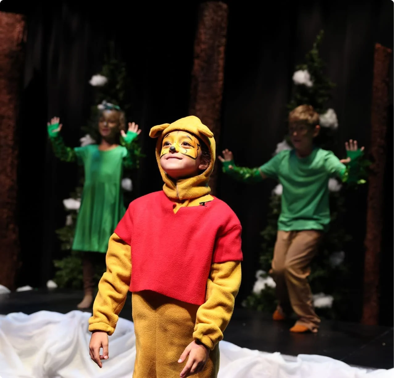 Children performing a play on stage dressed as animals, with one child in the foreground dressed as Winnie the Pooh and the others dressed as forest animals in the background.