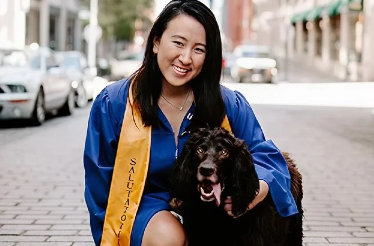 A young woman in a blue graduation gown and sash with the word 'Salutation' is smiling and kneeling beside a black Irish setter dog with a long coat on a city street.