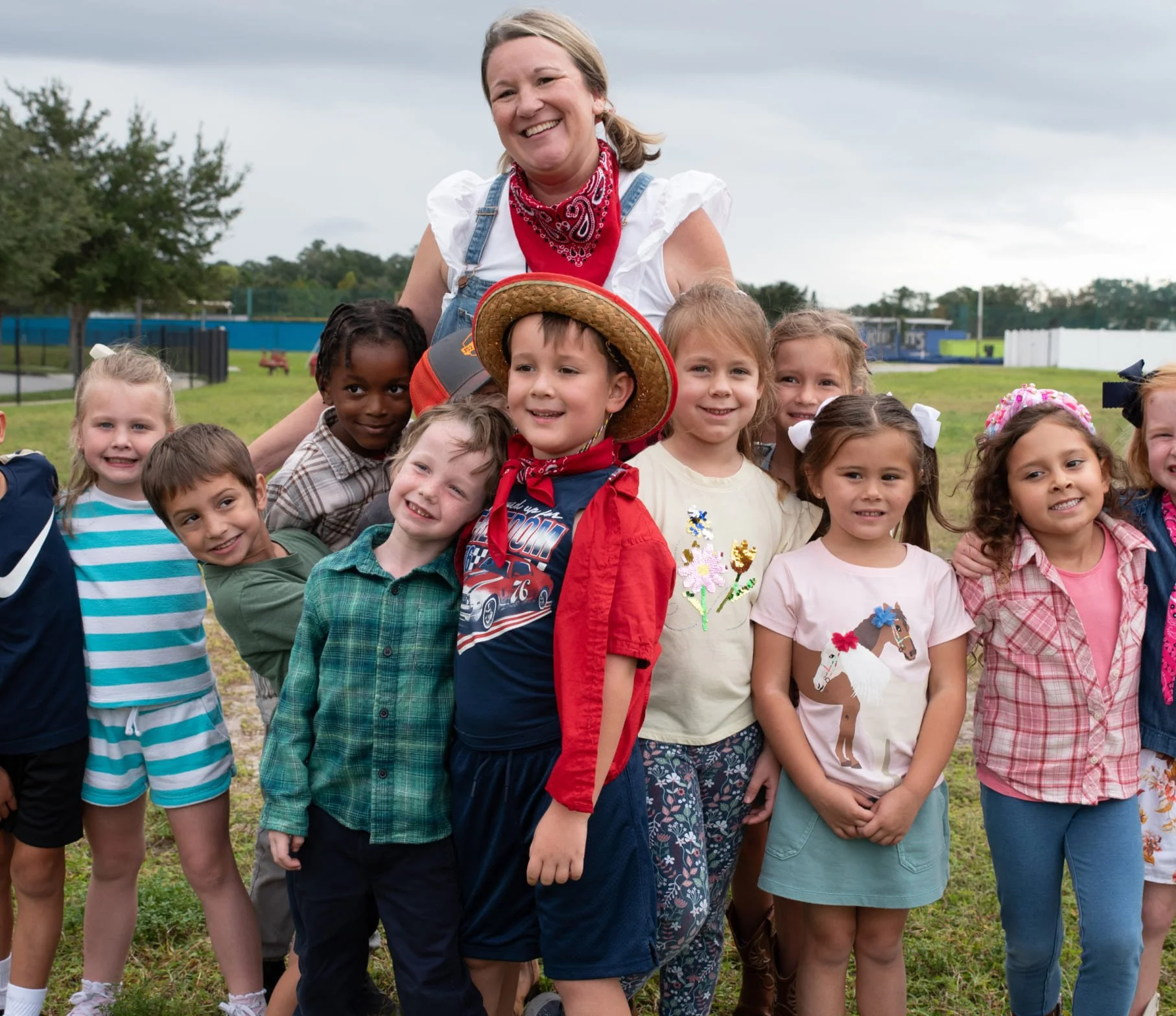 A group of children smiling and standing outdoors with a woman, possibly a teacher, in a park or playground setting.