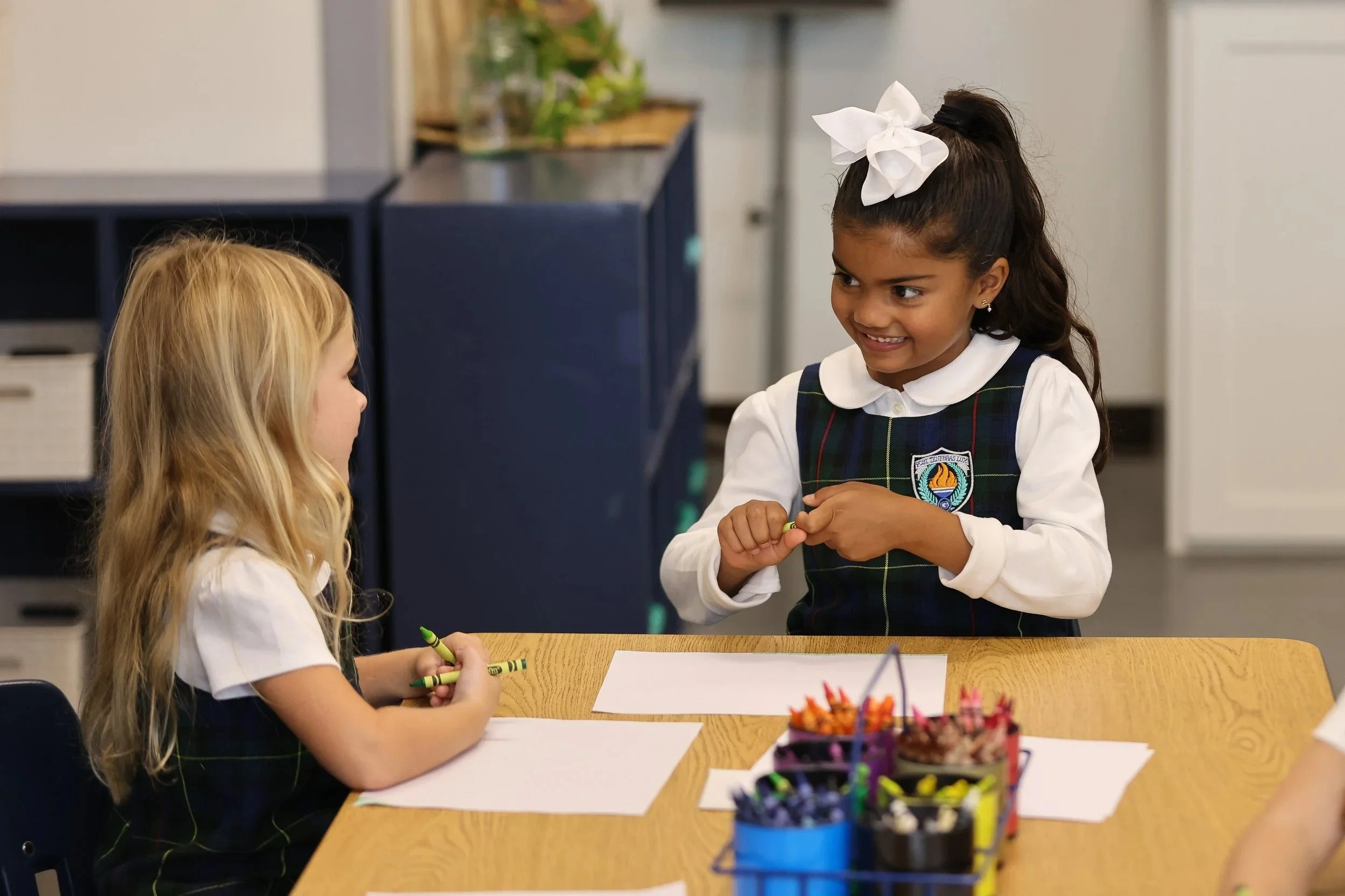 A classroom scene with two young girls in school uniforms talking and smiling.