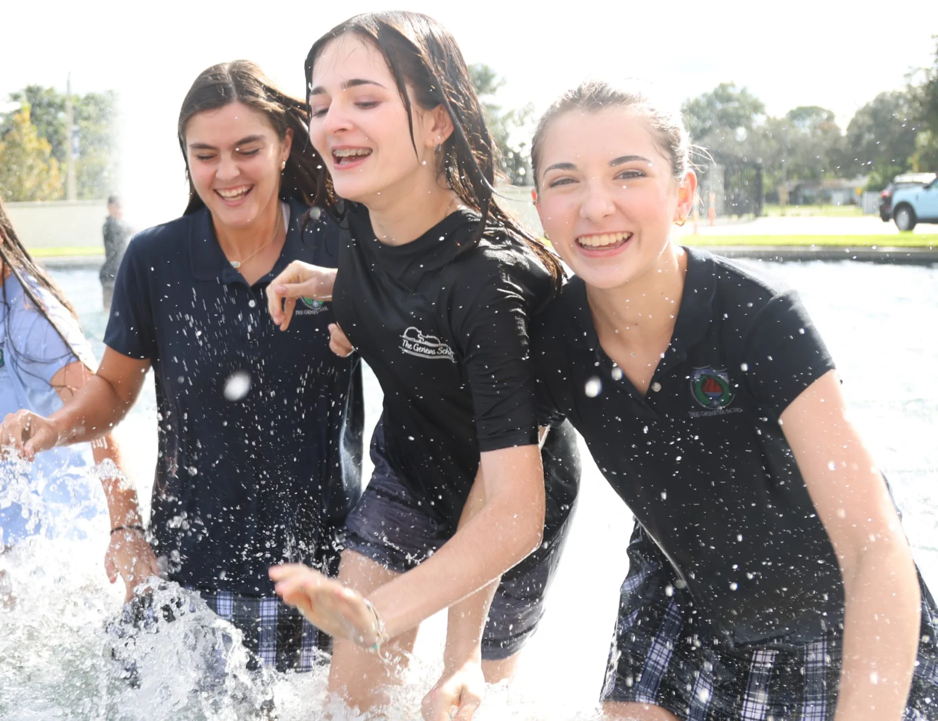Three students wearing Geneva's polo shirts and plaid shorts laughing and playing in water outdoors on a sunny day.