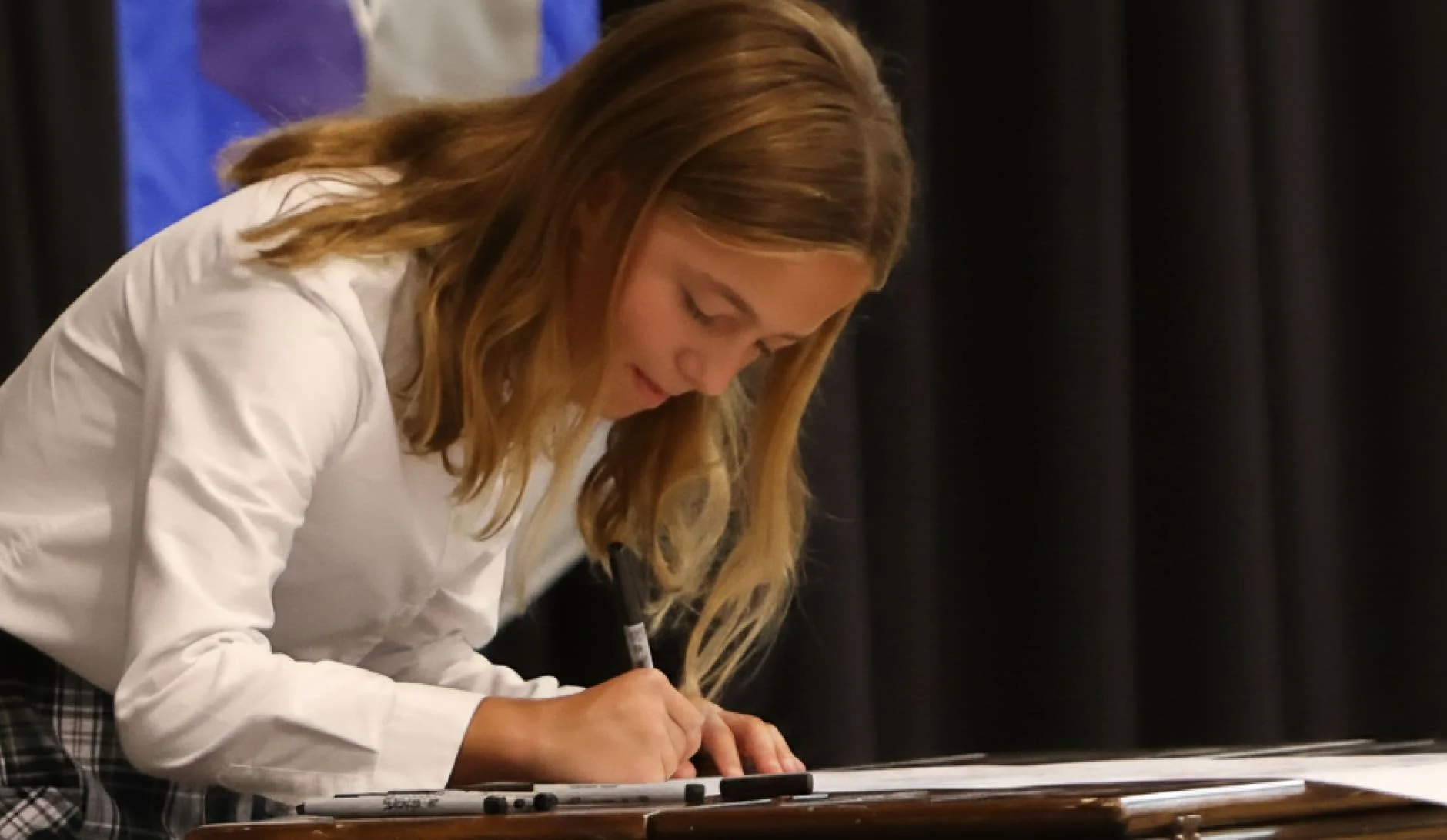 A student, wearing a white shirt, is signing a document on a table.