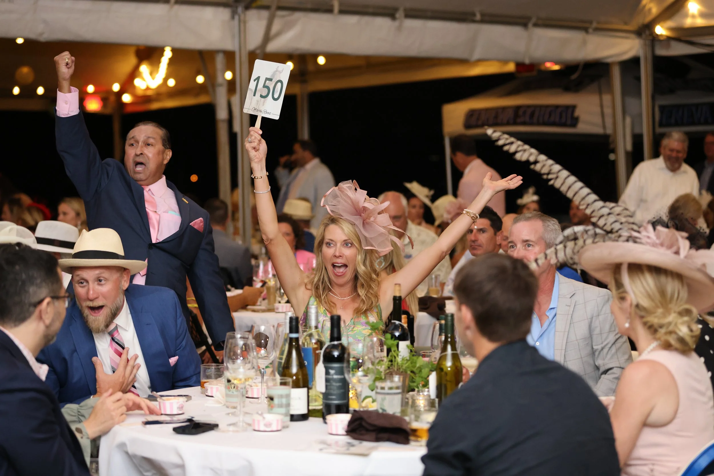 People celebrating at a dinner event, one woman holding a sign with the number 150, others raising their hands and smiling, decorated with string lights, wine bottles, and dinnerware on the table.