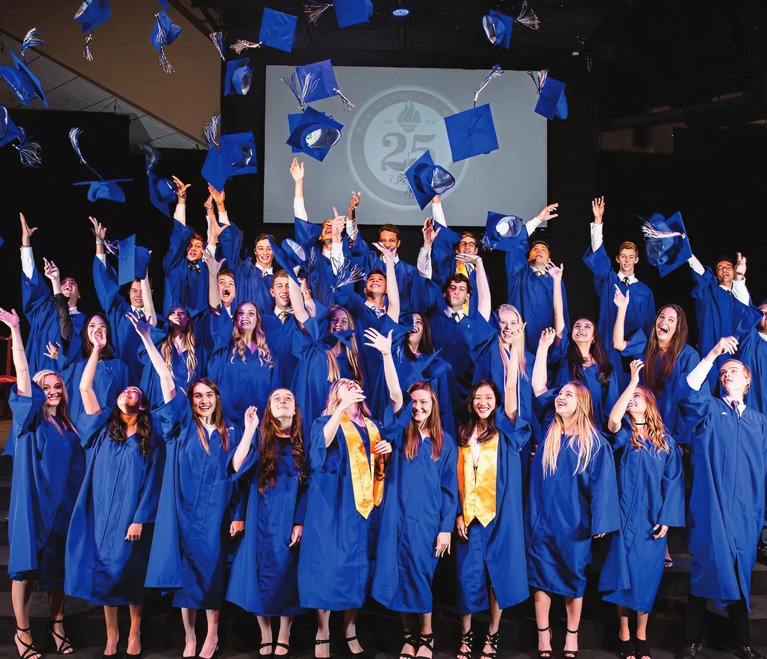 Group of graduates celebrating during a commencement ceremony, wearing blue caps and gowns, with some tossing caps into the air on stage.