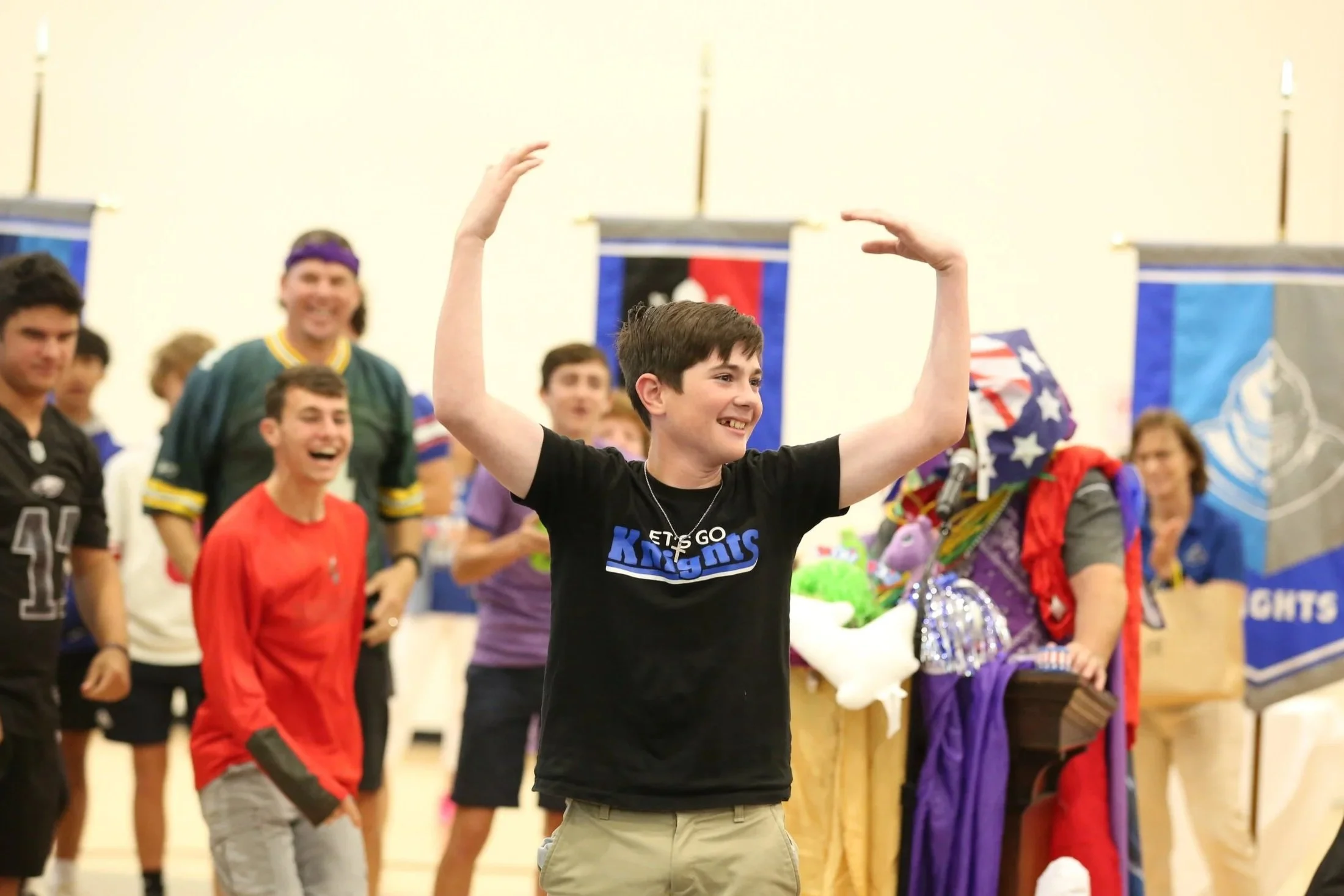 A group of young people at an indoor event, with one boy in the foreground smiling and raising his hands, and a woman in the background wearing colorful clothing and accessories.