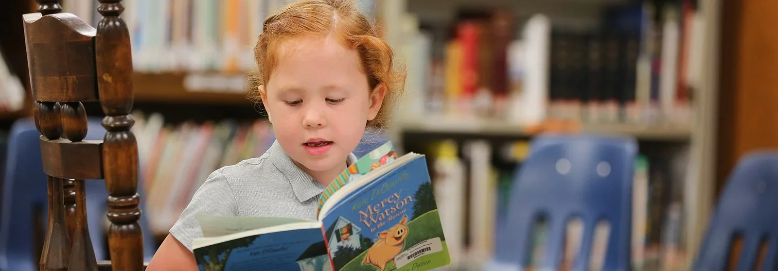 Young girl with curly red hair reading a children's book titled 'Mercy Watson' in a library.