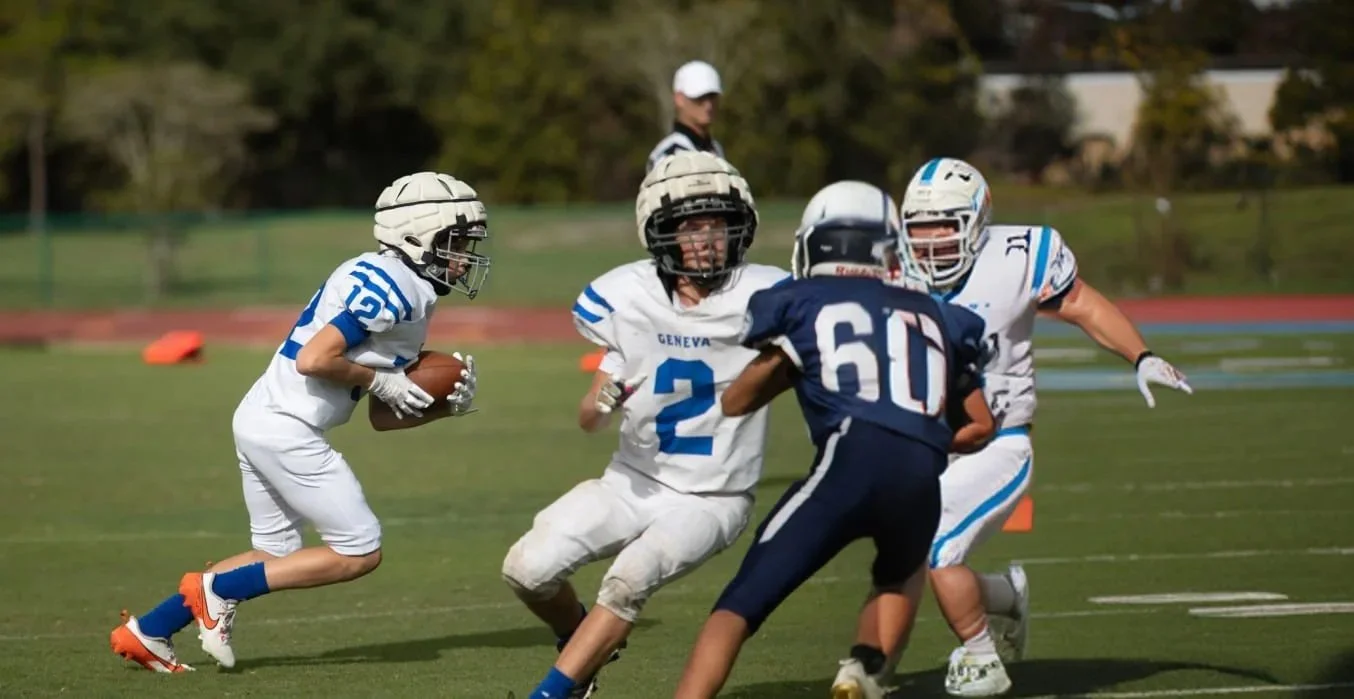 Young football players in white and blue uniforms playing on a field, with one player holding a football and others trying to block or tackle.