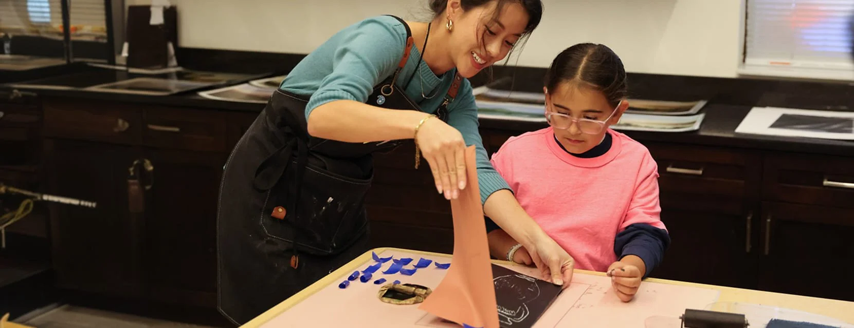A woman and a young girl working together on a creative project at a table, with the woman showing the girl how to create a paper sculpture or craft.