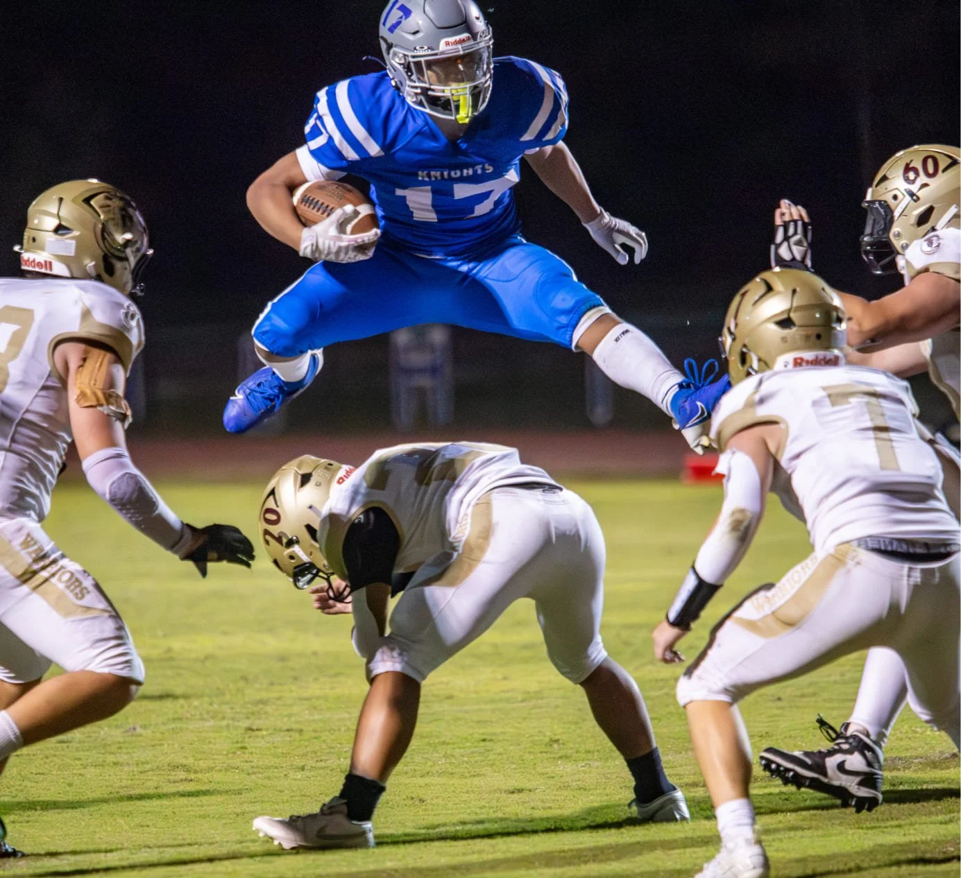 Football game at night with a player in blue uniform jumping over opponents in white and gold uniforms.